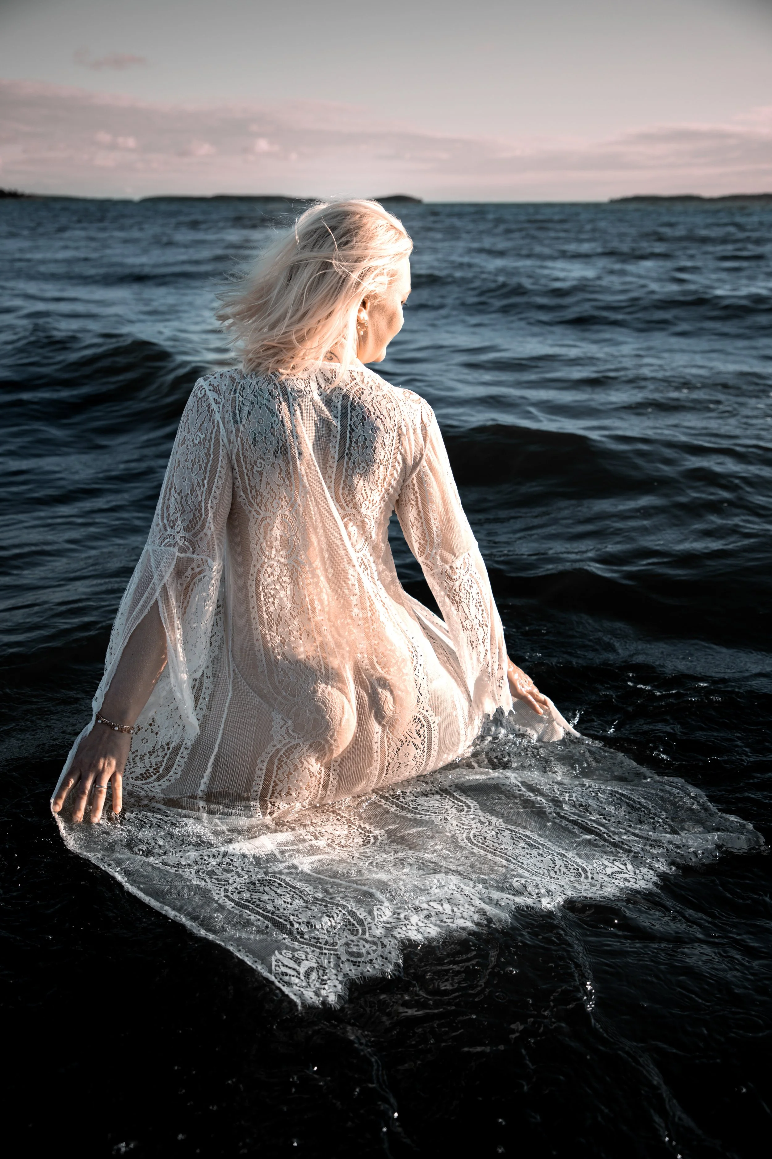A woman with blonde hair wearing a lace dress sitting in the ocean with her back to the camera, facing the horizon during sunset.