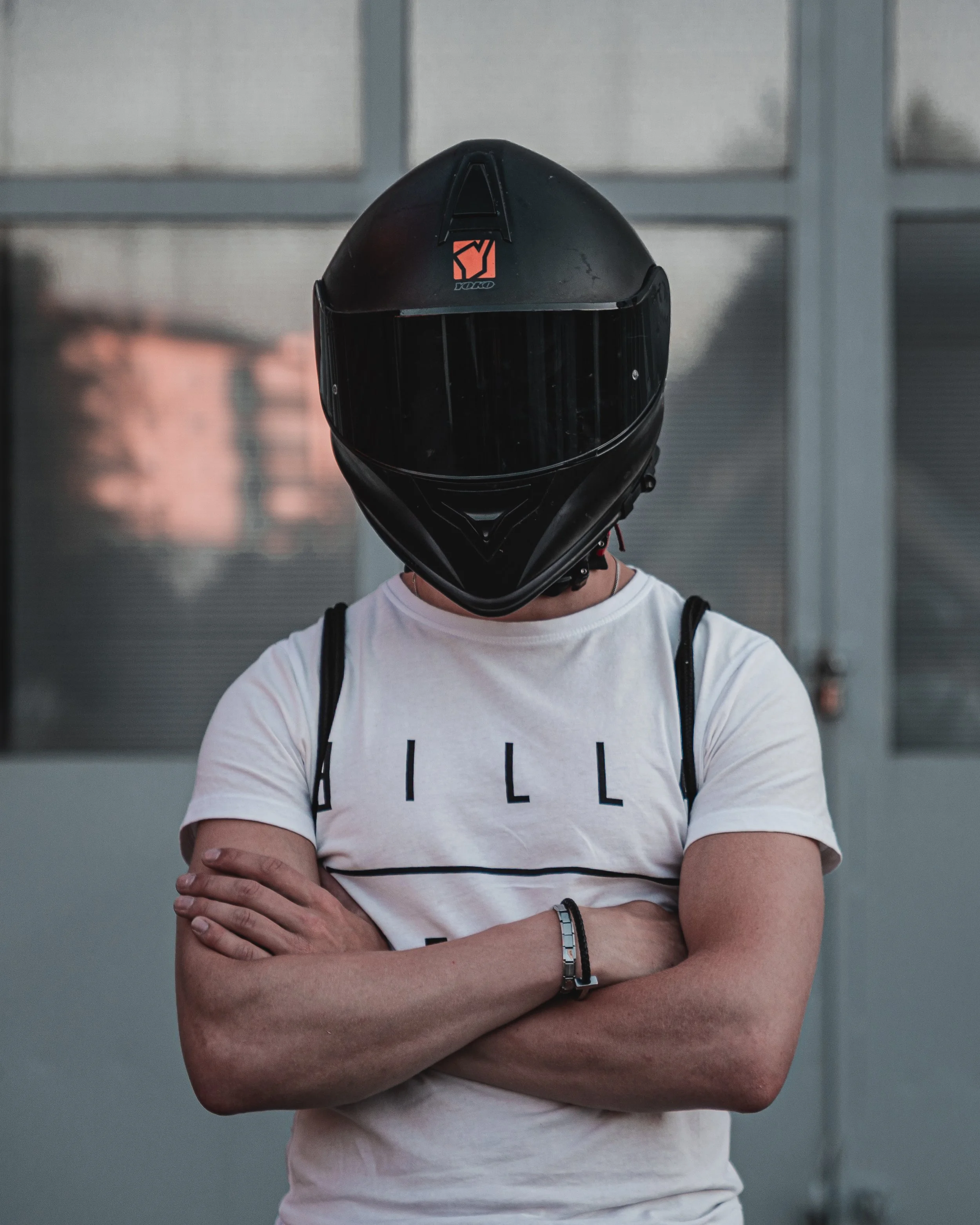 Person wearing a black motorcycle helmet with crossed arms, white T-shirt, and a black bracelet, standing in front of a metal fence.