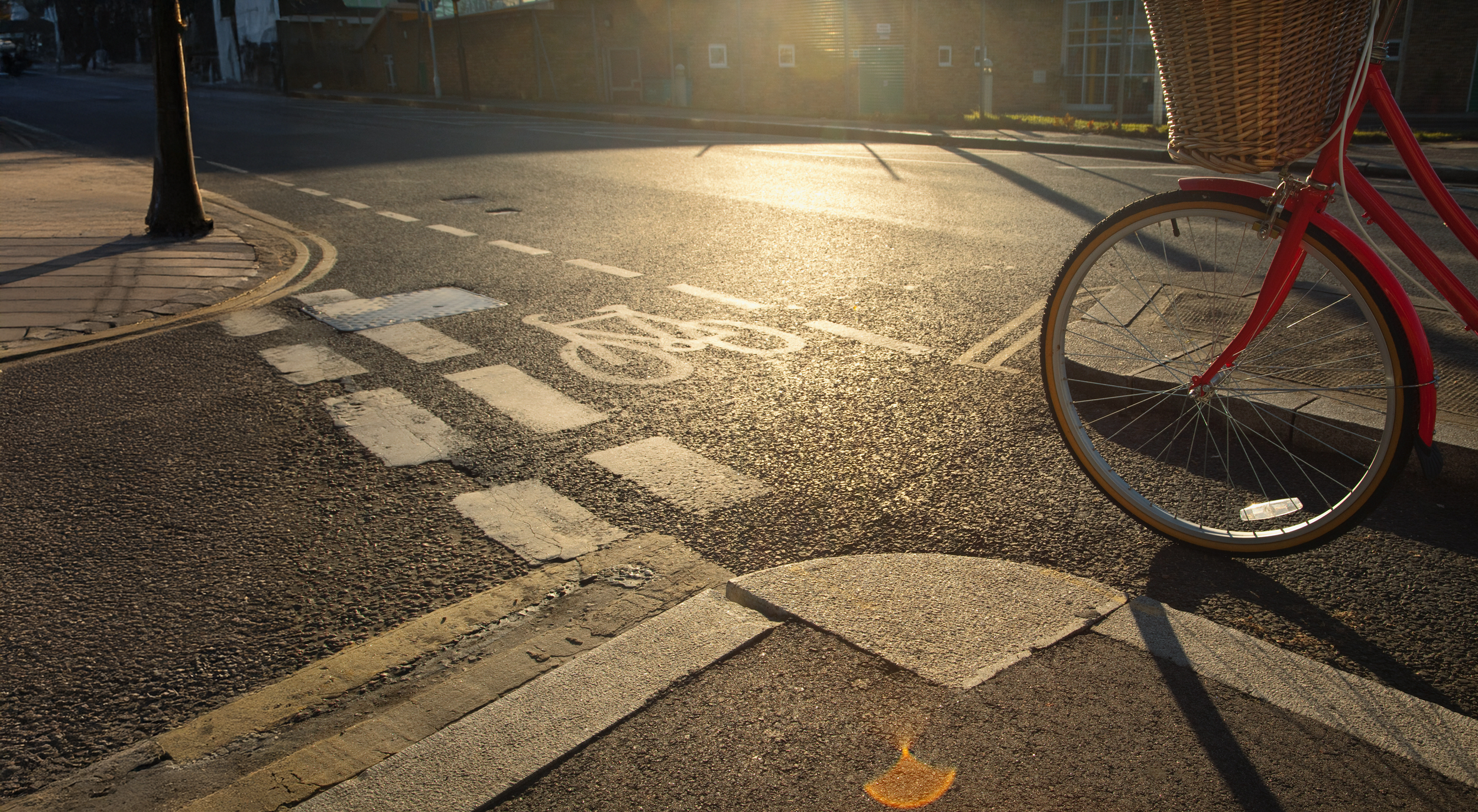 A red bicycle parked at a curbside with a basket on the front, on a city street during sunset, next to a bike lane and a crosswalk.