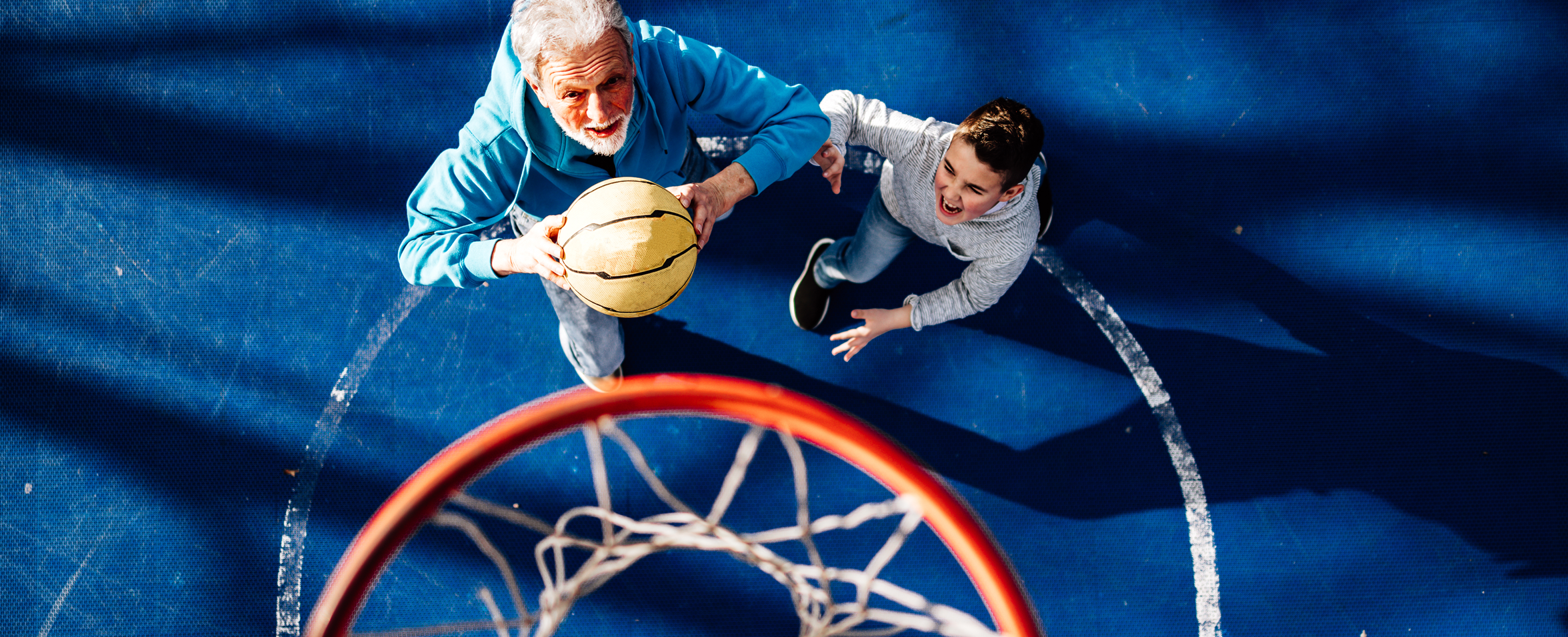 An older man and a young boy are playing basketball on an outdoor court, with the man holding the ball and both of them reaching towards the hoop.