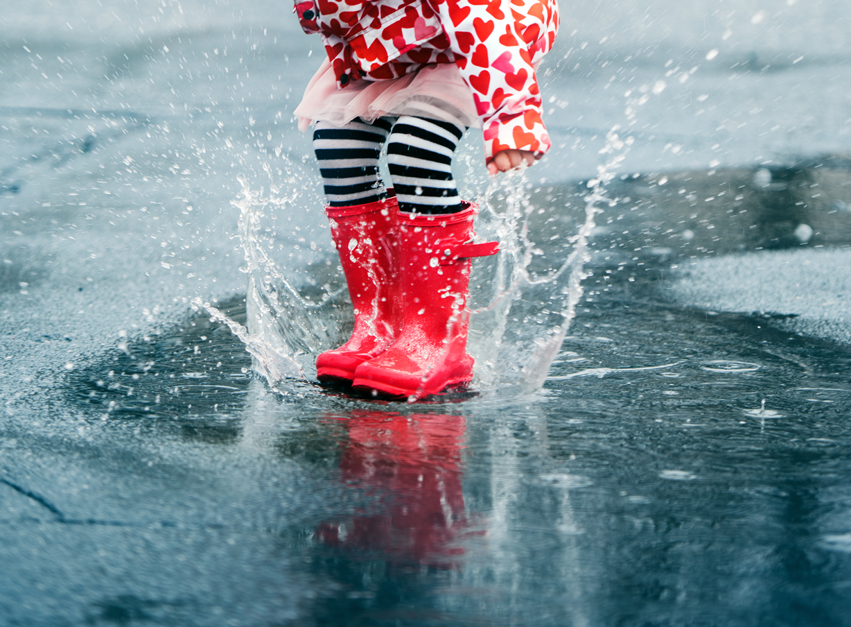 Child wearing red rain boots splashing in a puddle with water flying around.