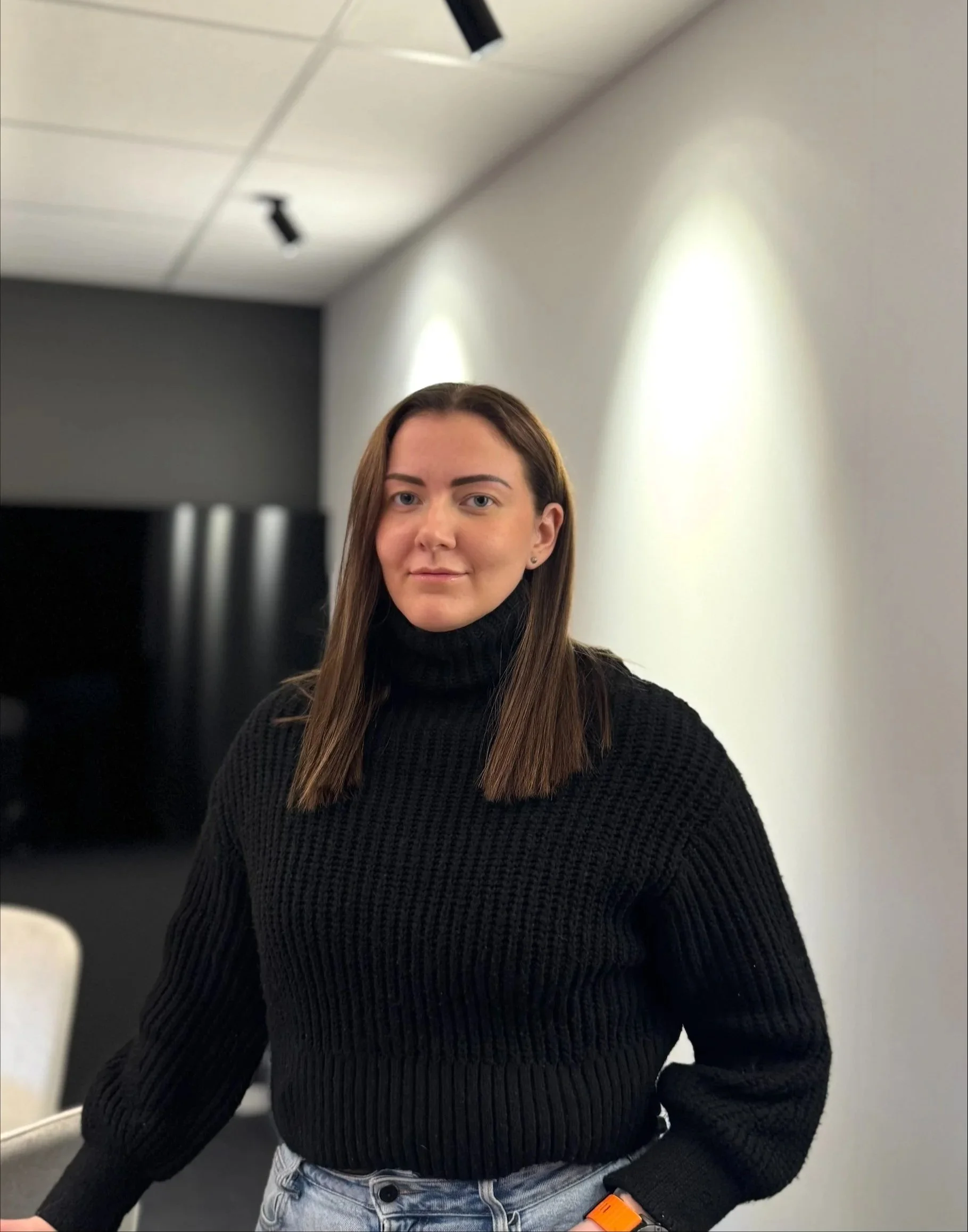 Et portrett av en ung kvinne med brunt langt hår iført en svart, ribbet genser og hvite bukser. Hun står i et rom med hvite vegger og tak, og det er taklys over hodet hennes.