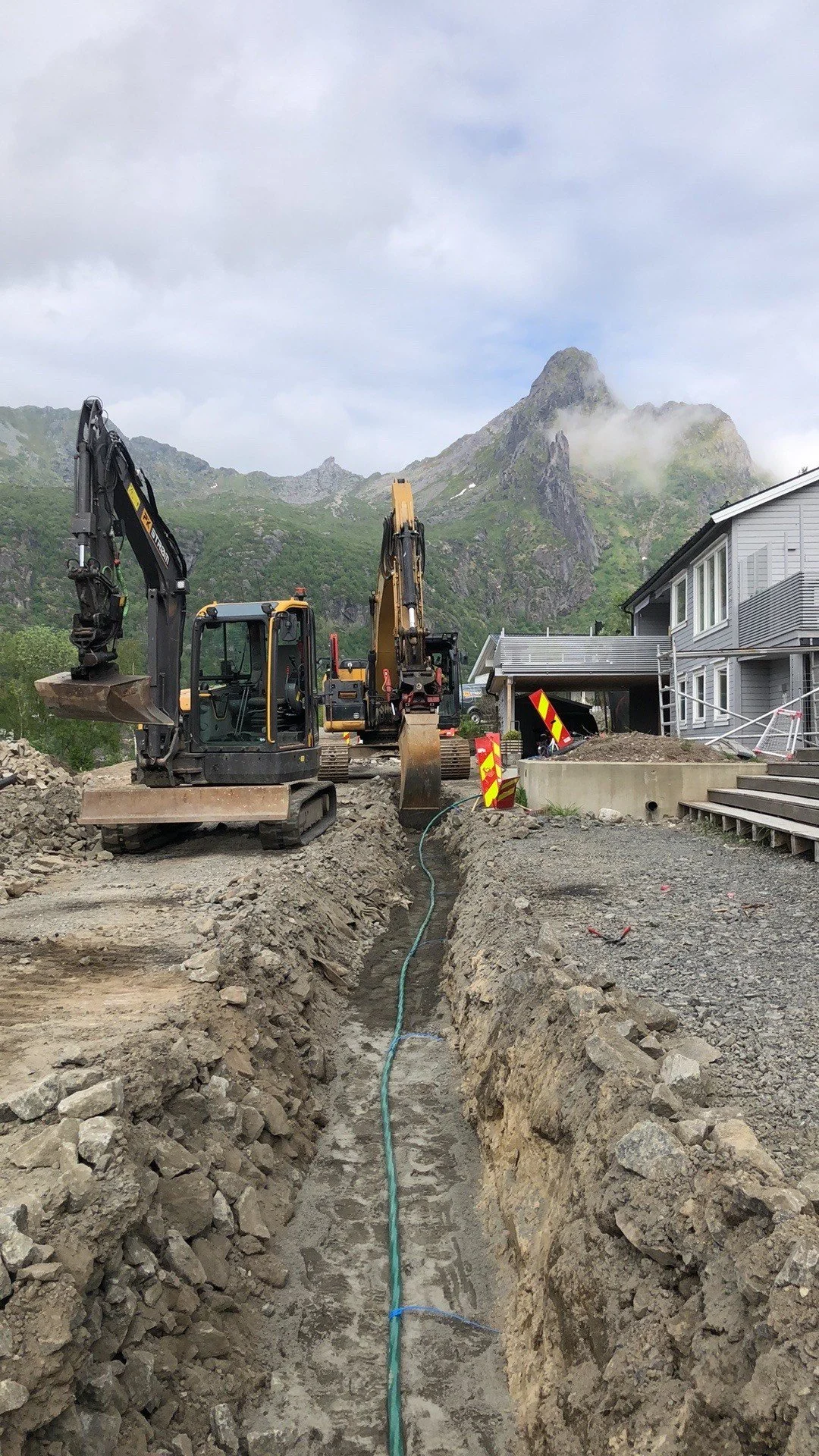 Byggearbeid med gravemaskiner som graver en asfalt før en bygning, omgitt av fjell og skyer.