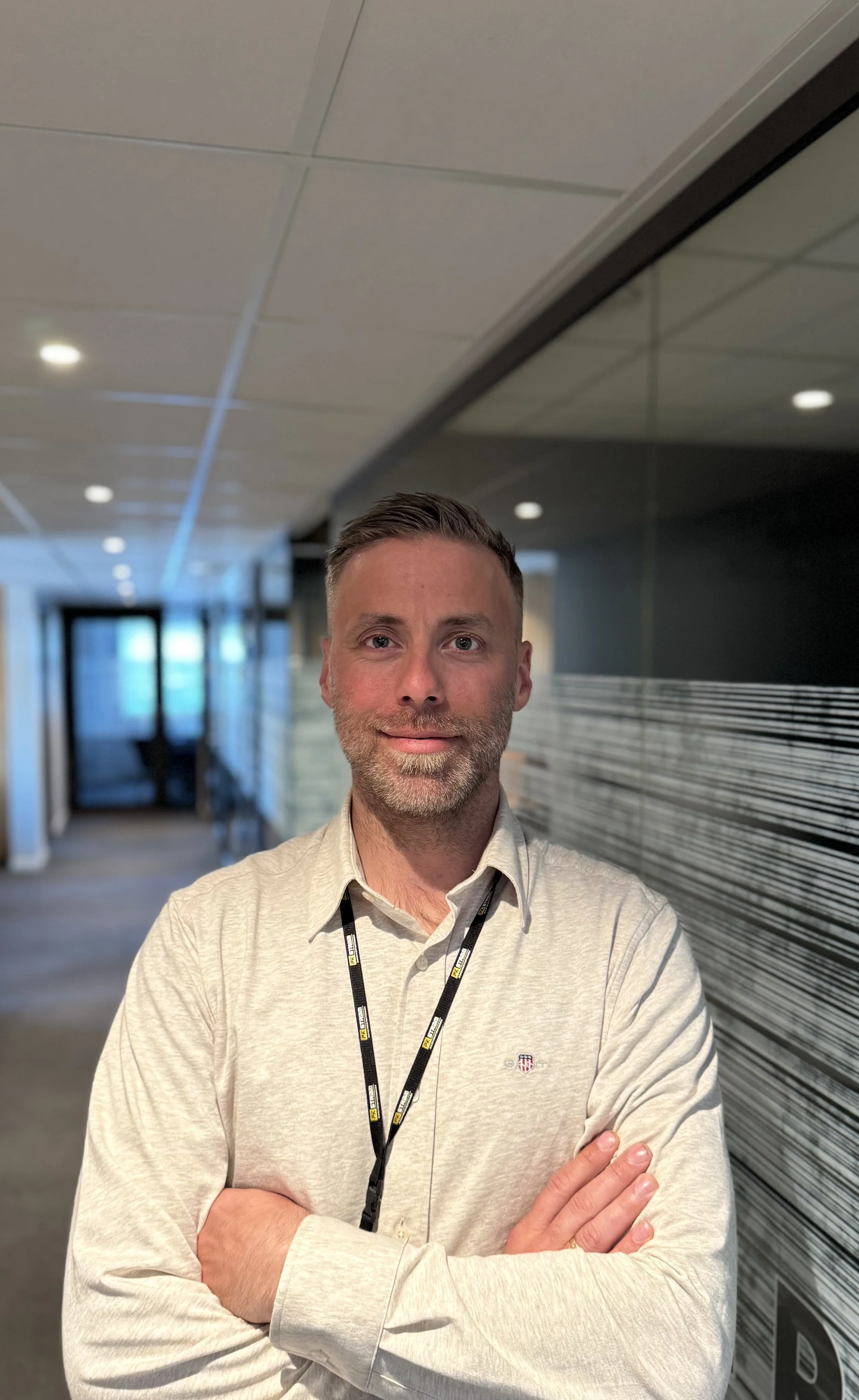 En mann med kort, lysebrunt hår og skjegg, iført lys skjorte og svart sele med logo, står med armene krysset foran en moderne kontorbygning. Bak ham er det en korridor med store vinduer og en vegg med horisontale striper.