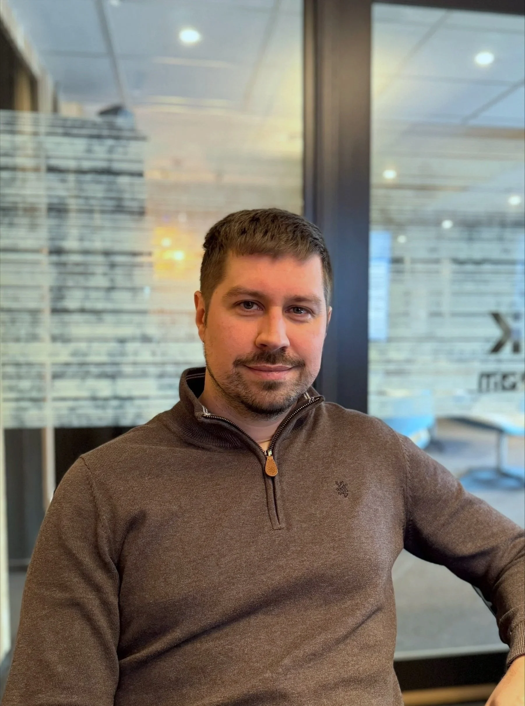 Et bilde av en mann med kort brunt hår og skjegg, iført en brun genser, sittende innendørs med en glassvegg i bakgrunnen.