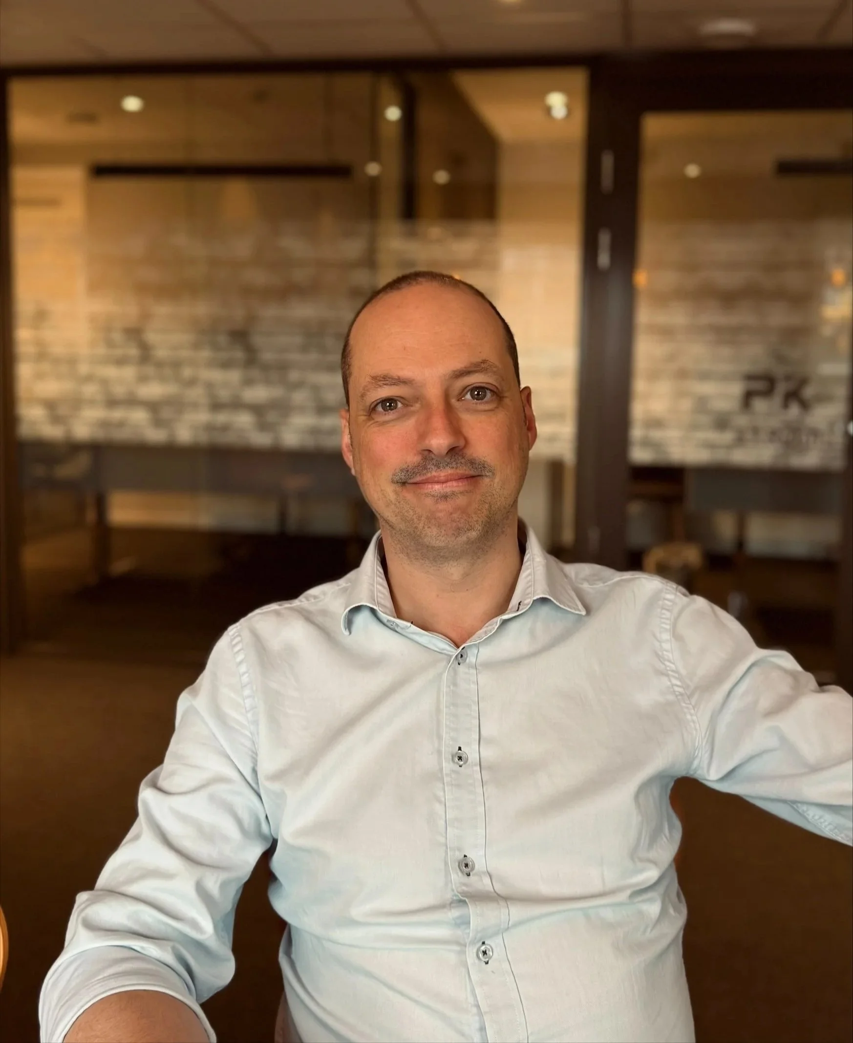 Liten mann med kort hår og skjegg smiler til kameraet i et rom med tre vegger i bakgrunnen, bunnen av bildet viser en person iført hvit skjorte.