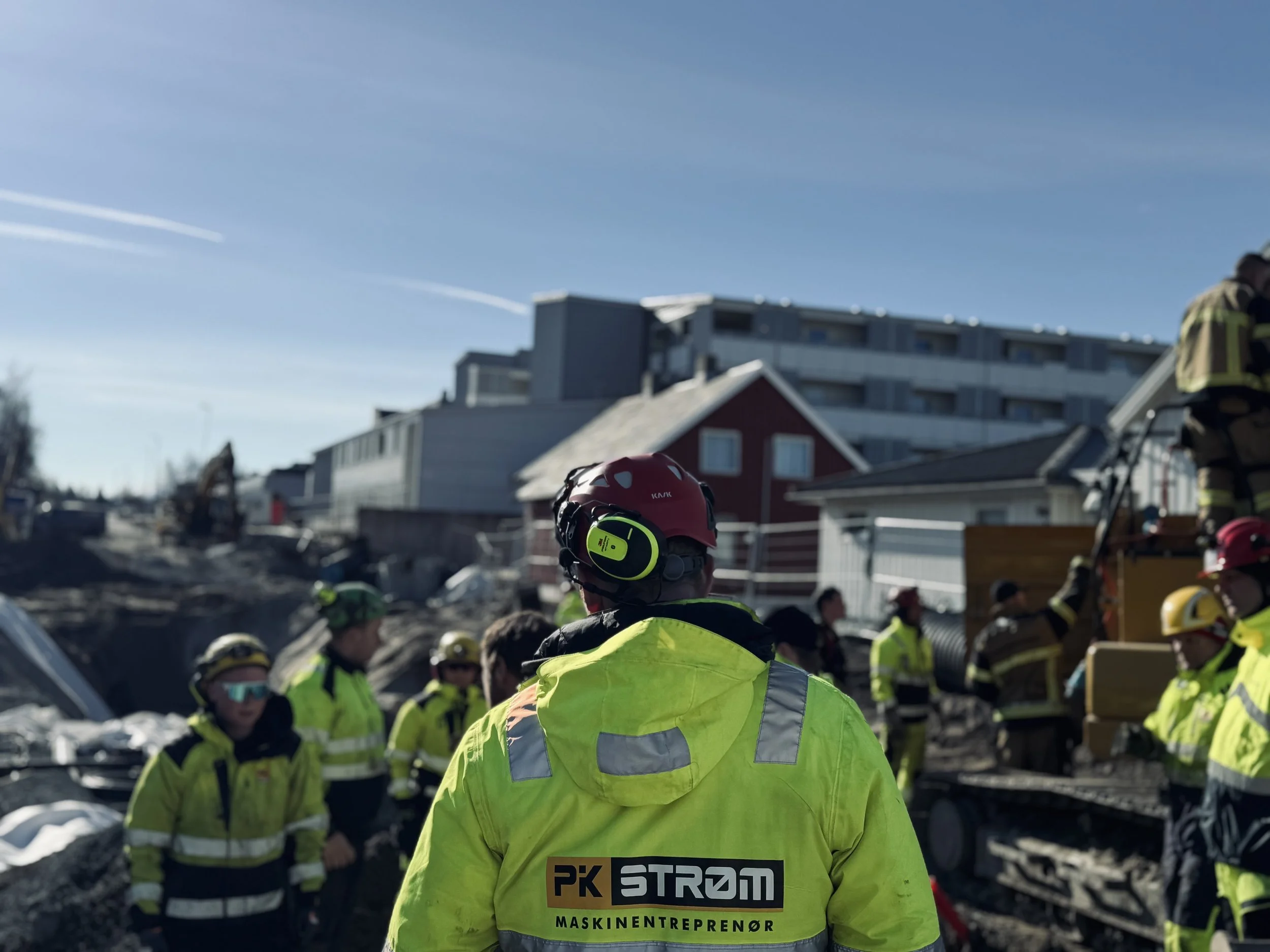 Grupp mennesker i synlige jakker, noen med hjelm, utenfor en bygning i solen, anleggsarbeid, med flere mennesker i bakgrunnen. PK strøm