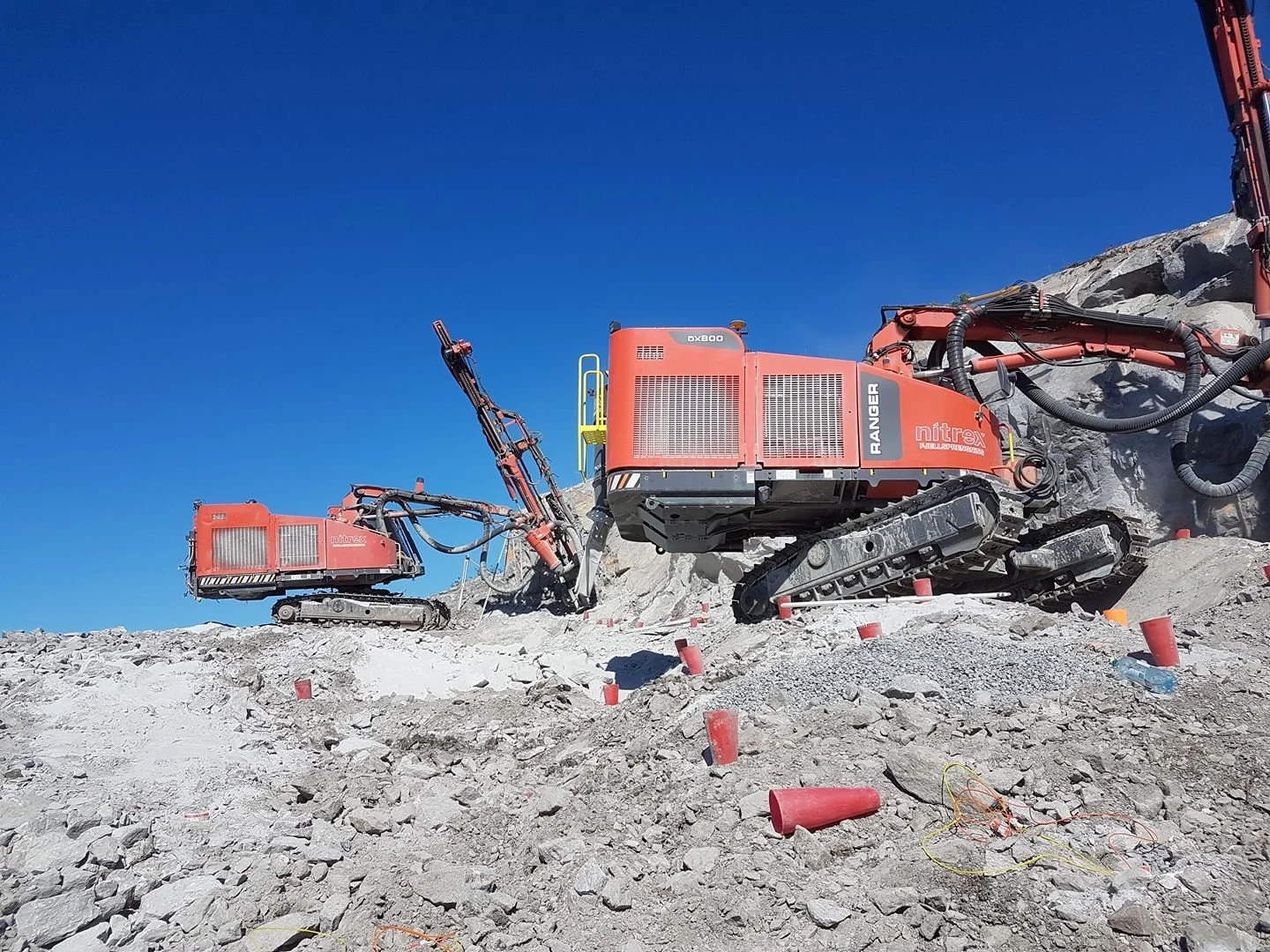 To store gravemaskiner, et terreng av grus og steiner, blå himmel.