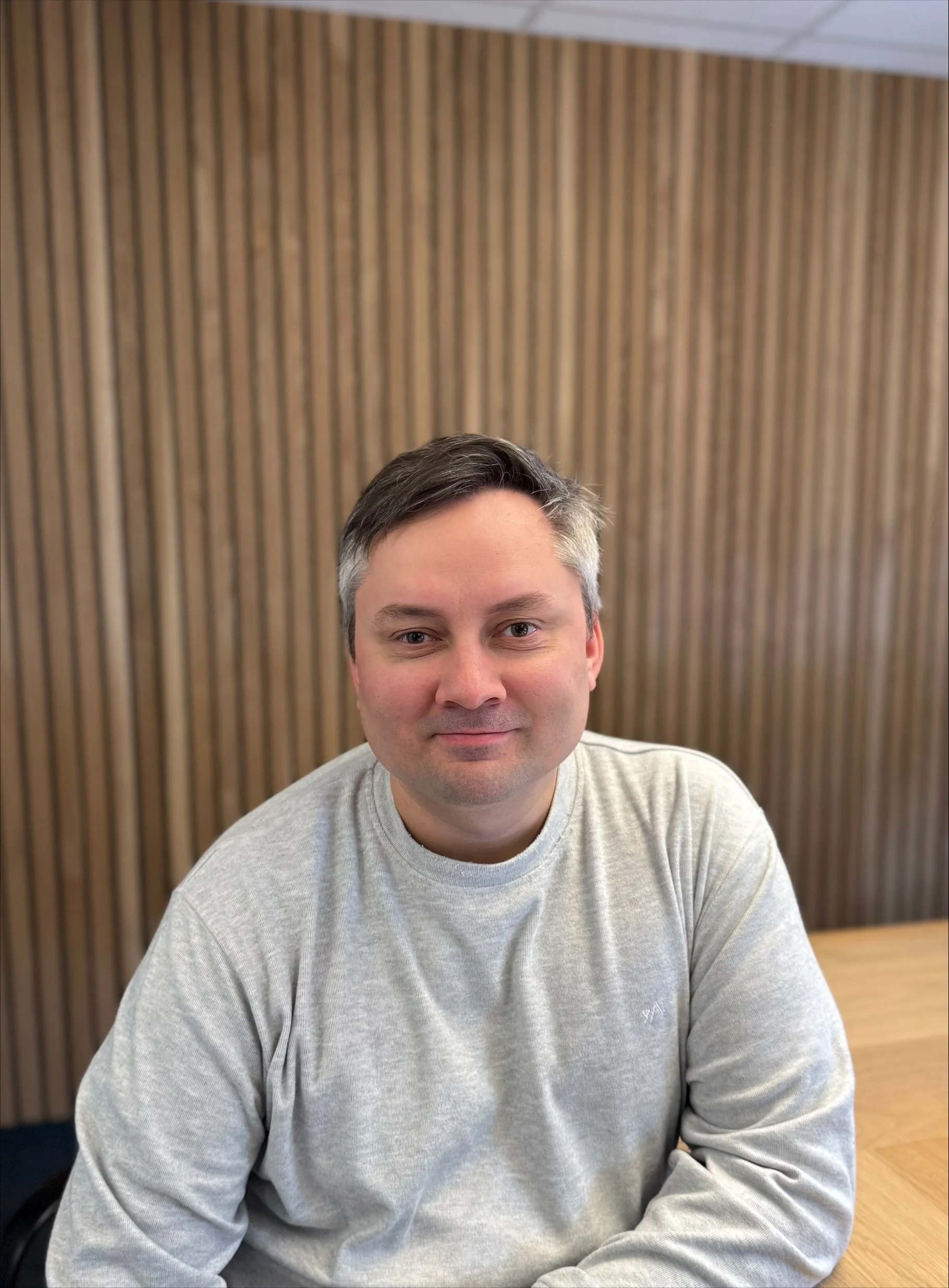 En mann med kort, mørk og grått hår, iført en lys grå genser, sitter ved et bord foran en vegg med treplanker.