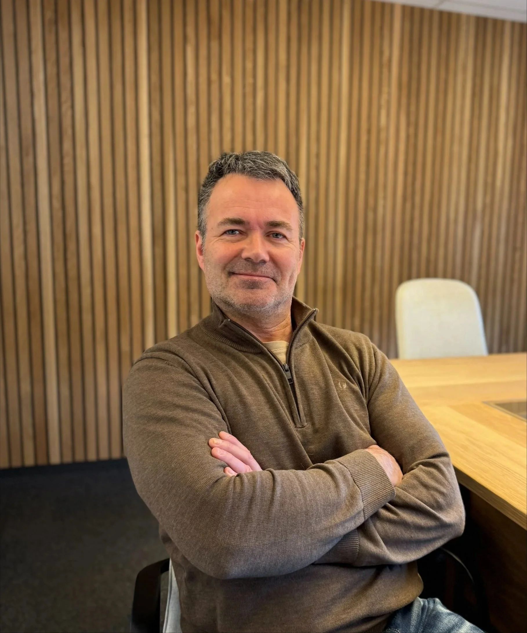 En mann med mørkt hår og skjegg smiler, iført en brun genser, sitter ved et bord i et rom med panelvegg og lampe. Han har armene krysset.