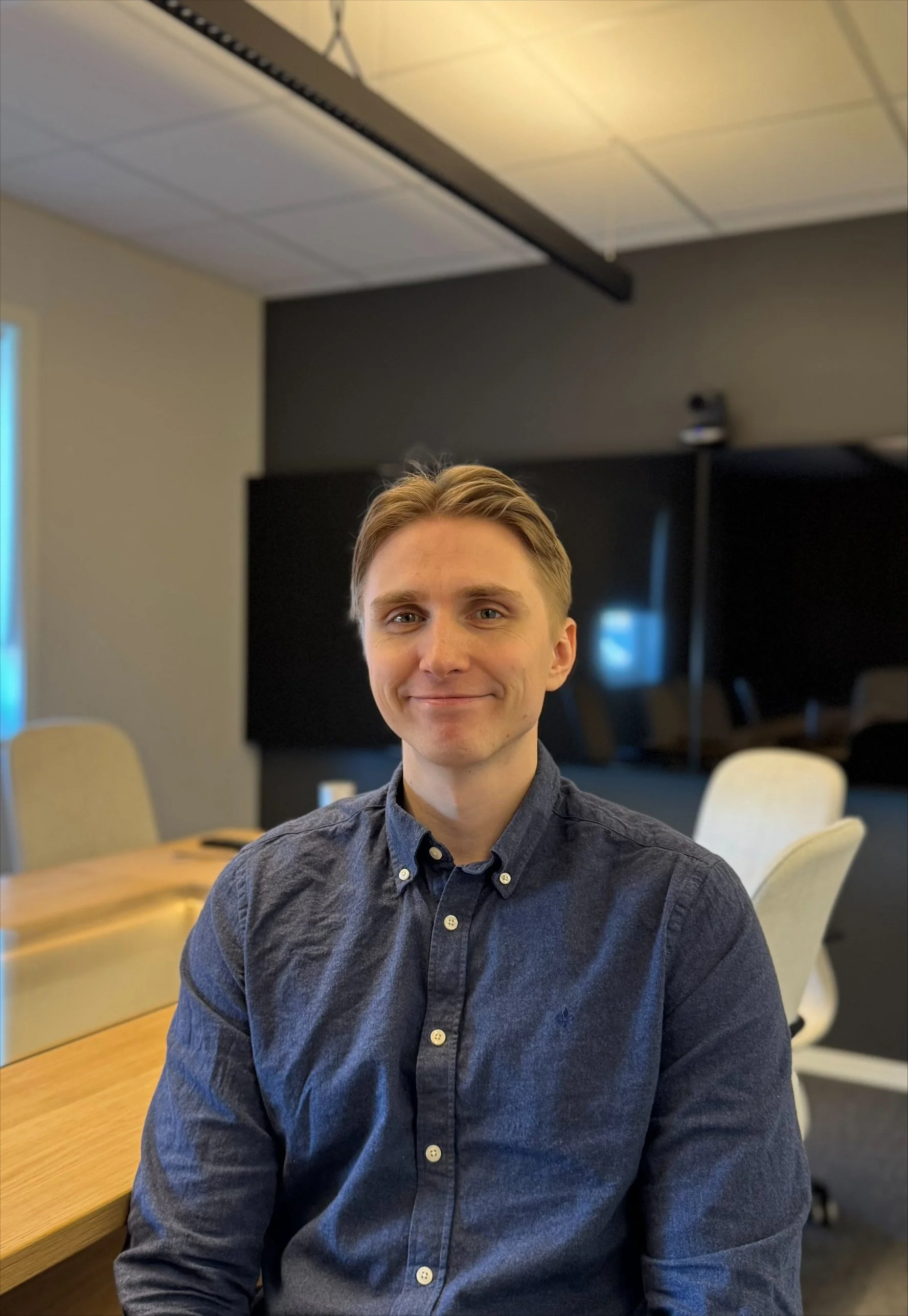 Et smilende ungt mann med lysebrunt hår iført en mørkeblå knappestilt skjorte, sitter ved et rundt trebenet bord i et møtelokale.