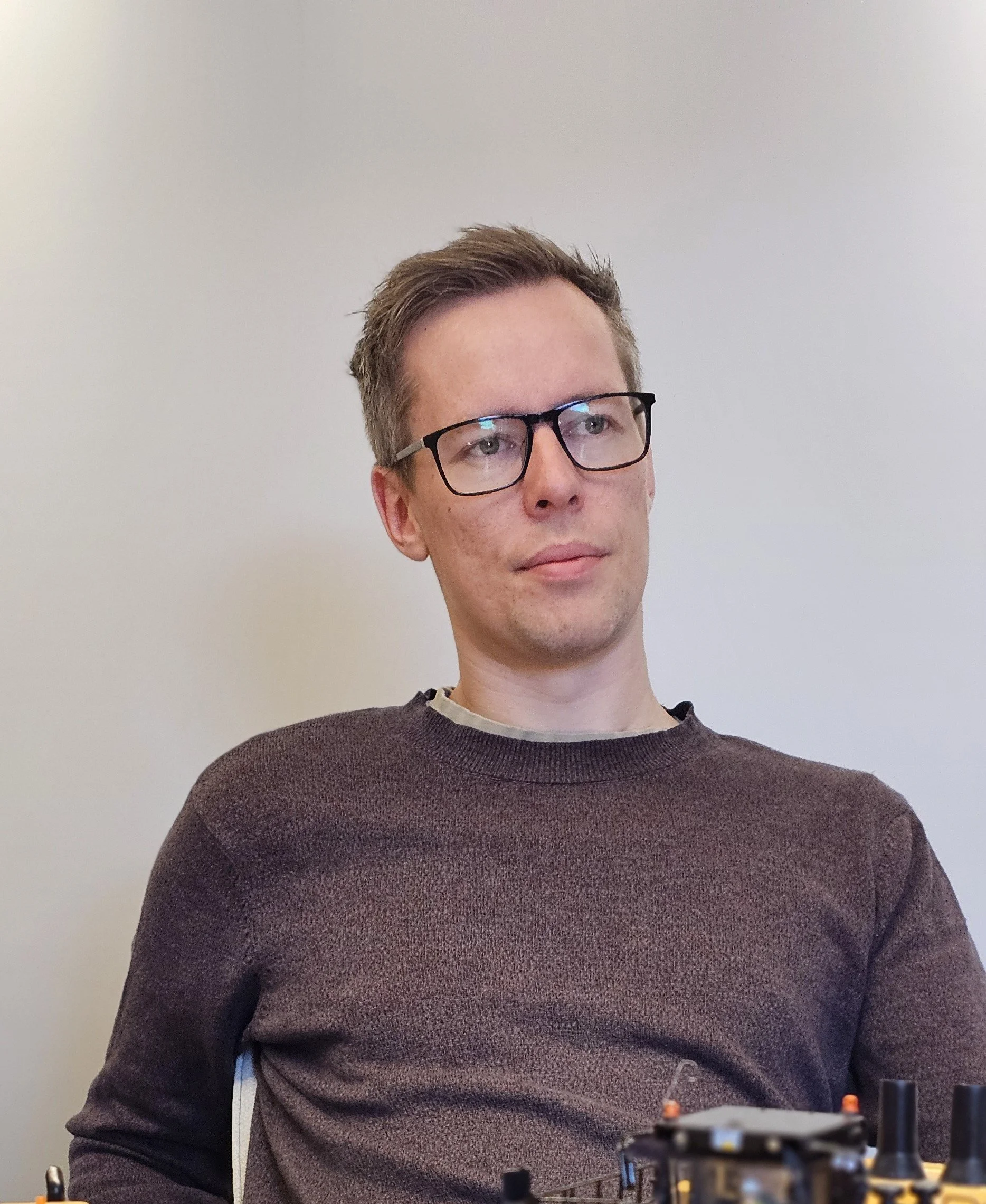 Dette er et portrett av en mann foran en hvit vegg. Han har brunt, kort hår og bærer briller. Han har en rolig utstråling.