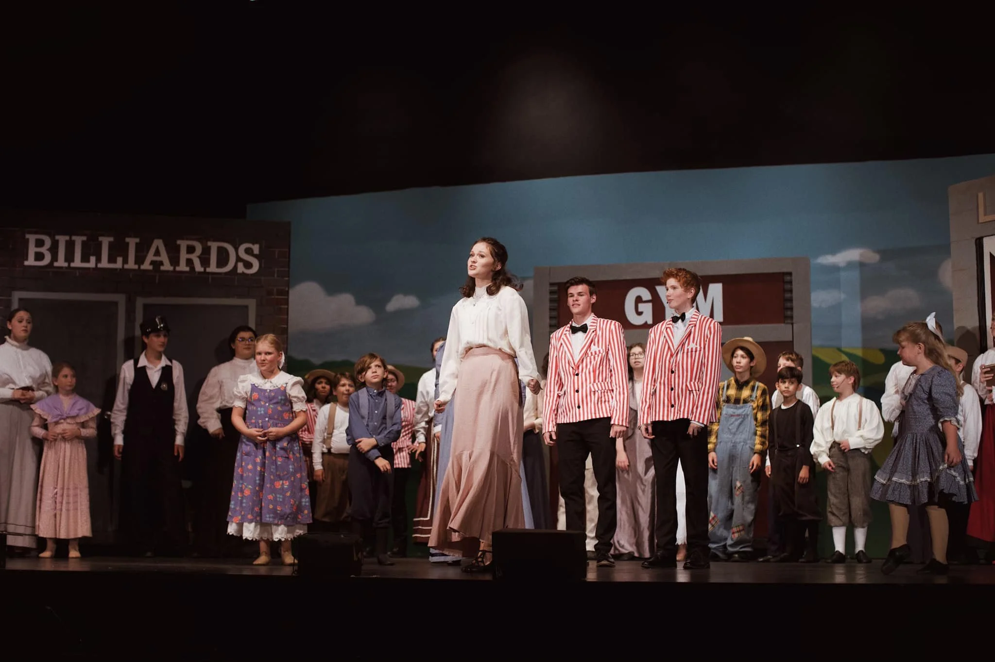 Stage performance with children in period costumes, including a woman in a white blouse and pink skirt, and two boys in red and white striped jackets, on a set with signs reading 'BILLIARDS' and 'GYM'.