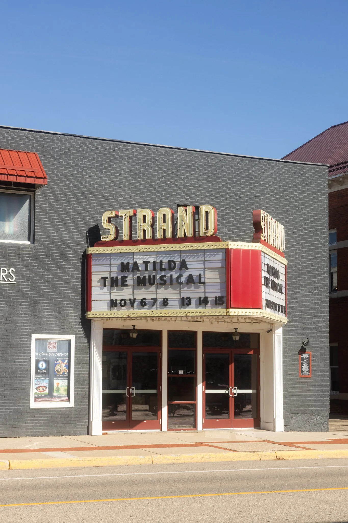 A photograph of The Strand Theater of Alma Michigan. It is a vertical view of the front doors and marquee in front of a clear blue sky.