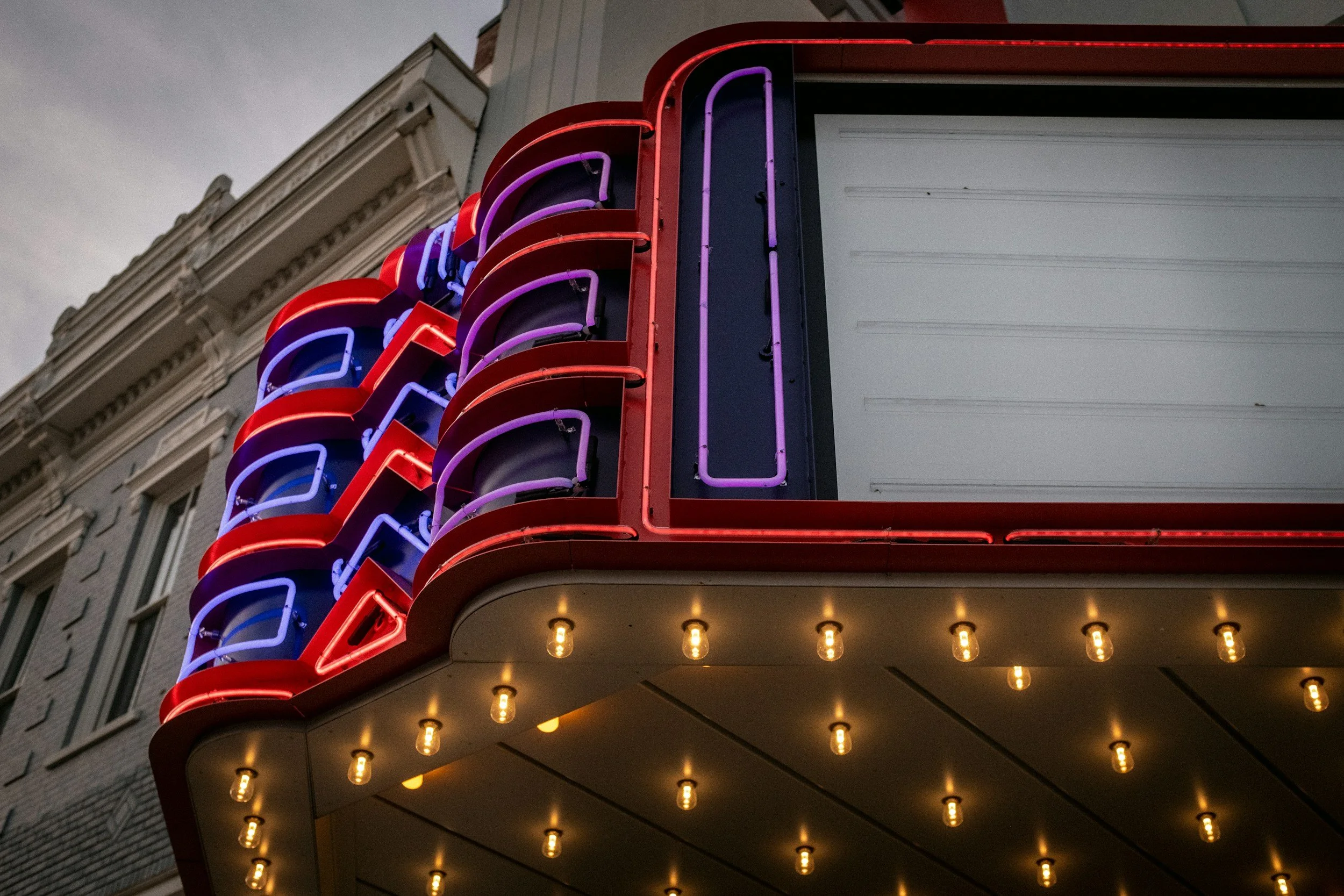 Neon sign on building with red, purple, and blue lights, the bottom part with warm yellow bulbs, against a cloudy sky and older building architecture.