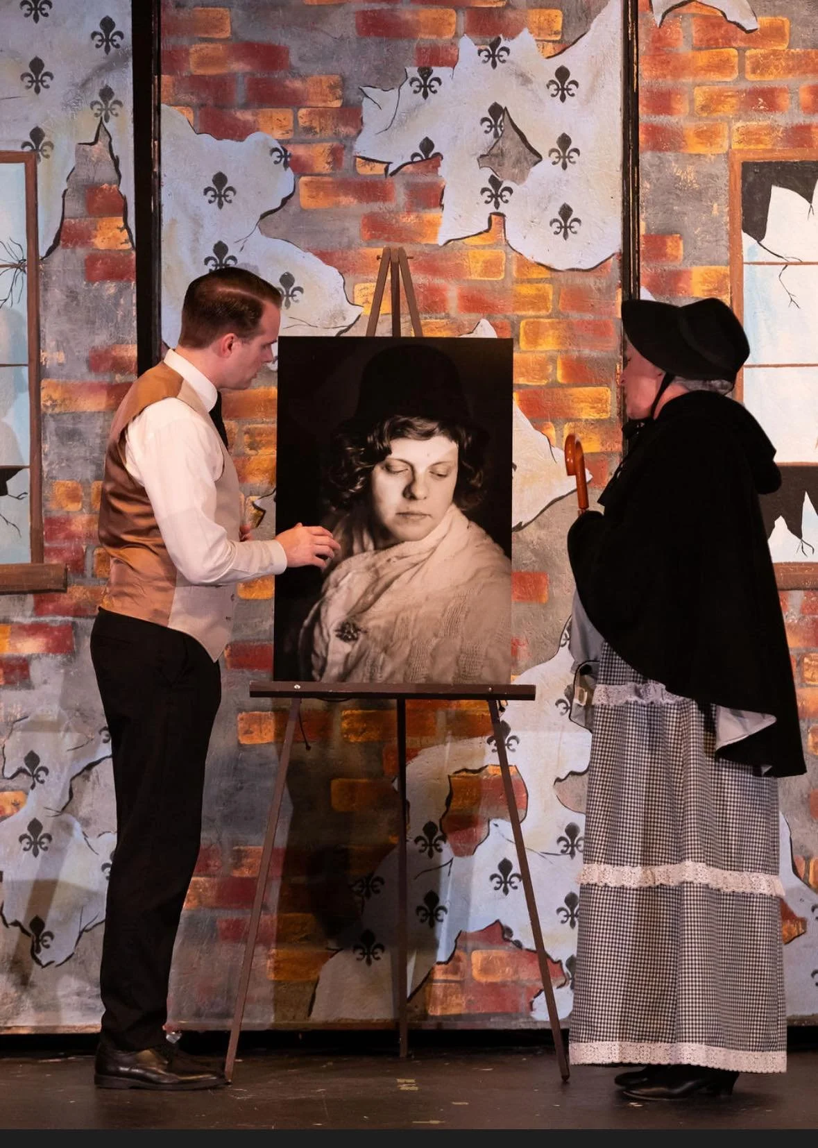 Two individuals dressed in vintage clothing looking at a black and white portrait of a woman on an easel in front of a distressed brick wall.