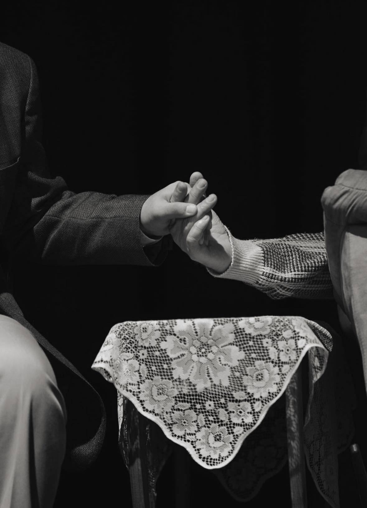 Two people holding hands, one wearing a suit and the other wearing a sweater, with a lace tablecloth on the table in front of them.