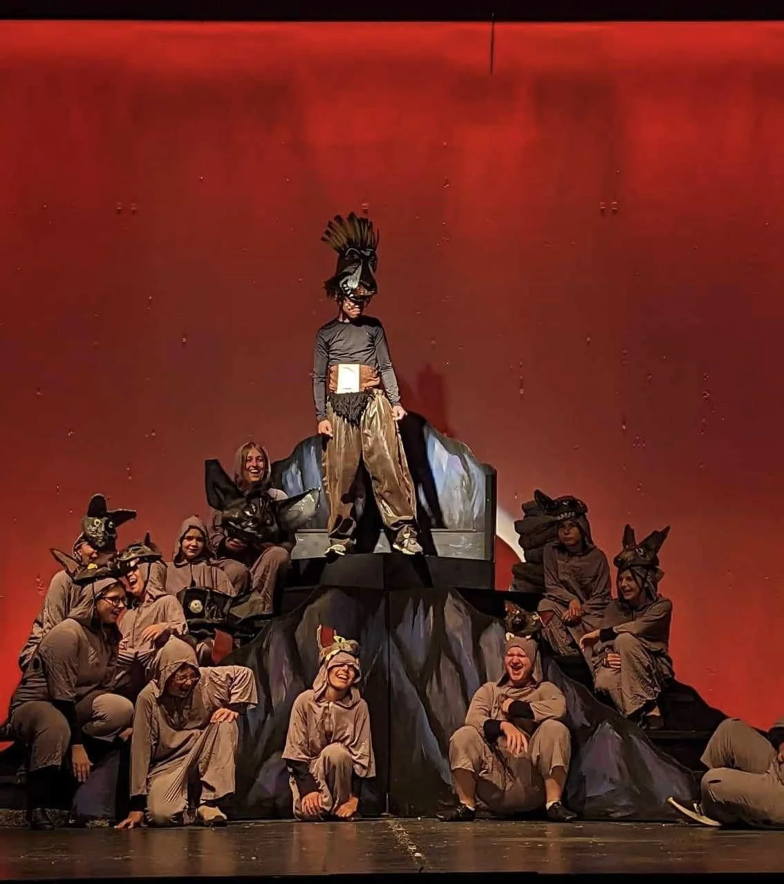 Children dressed in animal costumes participating in a stage play on a mountain-shaped set with a red background.
