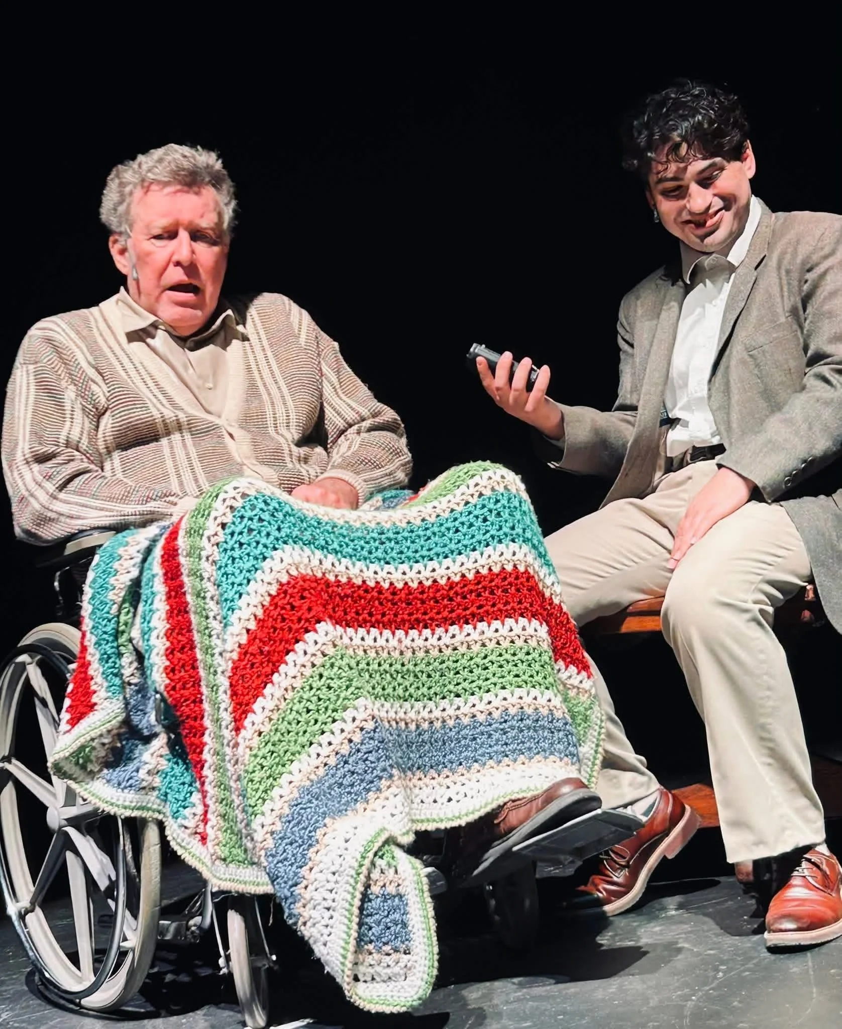Two men, one in a wheelchair and the other sitting beside, share a moment on stage. The man in the wheelchair is wrapped in a large, multicolored crocheted blanket, and the man beside him is holding a small object, possibly a remote or phone, smiling.
