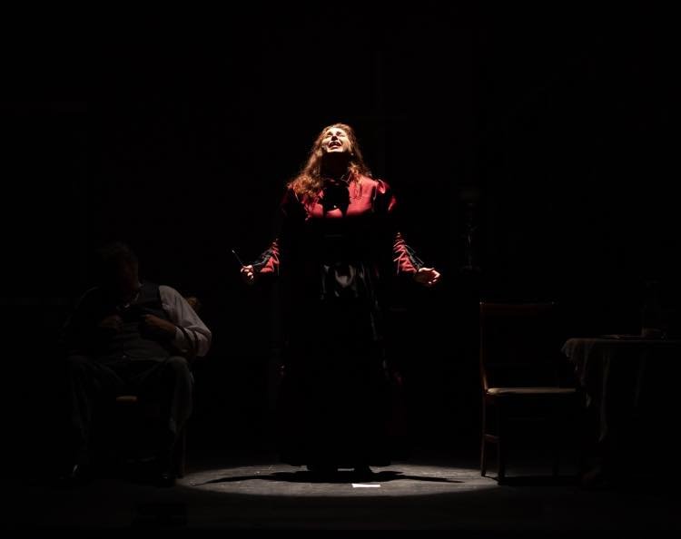 A woman with long hair wearing a red jacket is standing on a stage in a spotlight, possibly singing or speaking, with two seated individuals in the background in a dark setting.