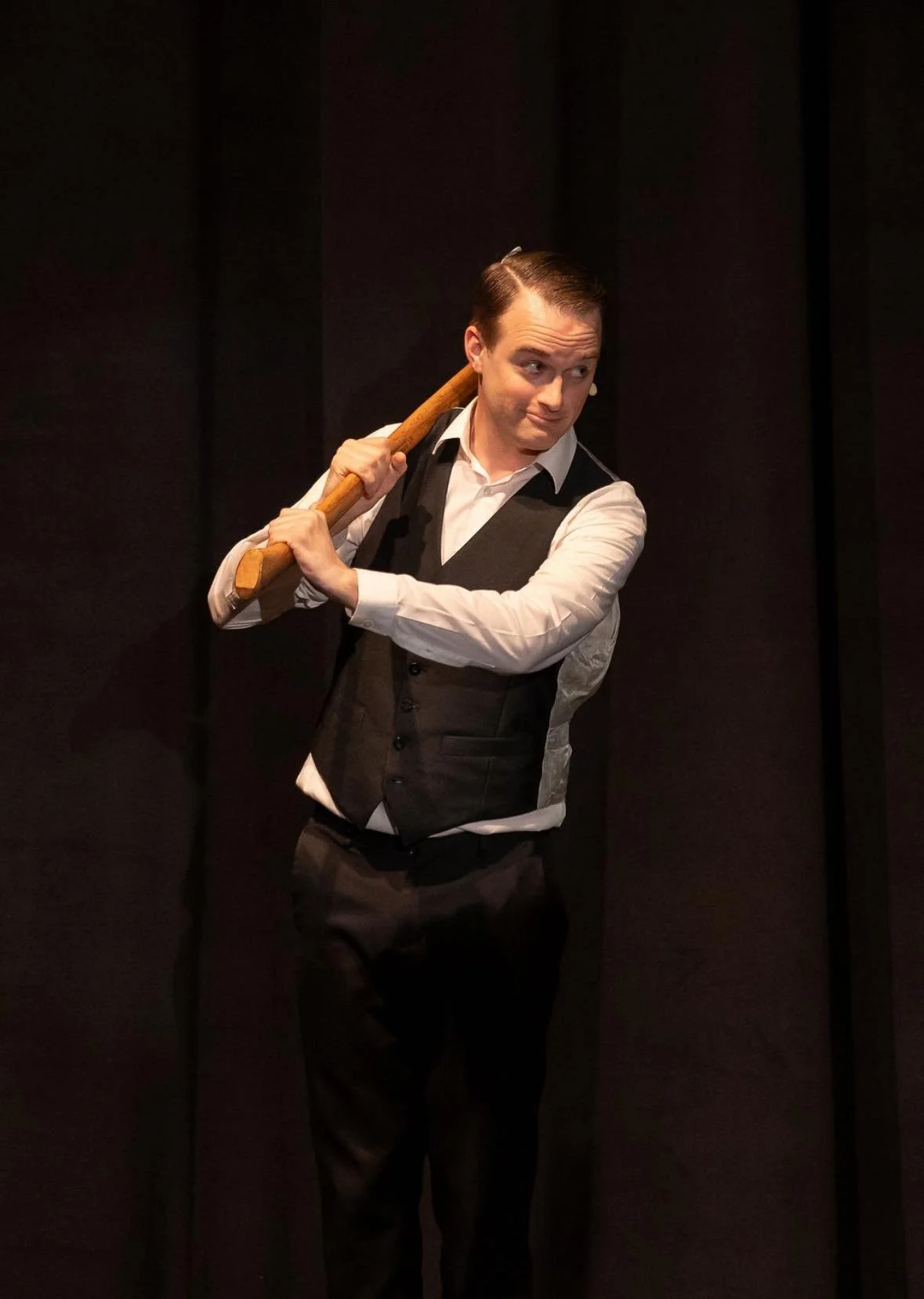 Performer in formal attire holding a wooden baton, standing against a dark background.
