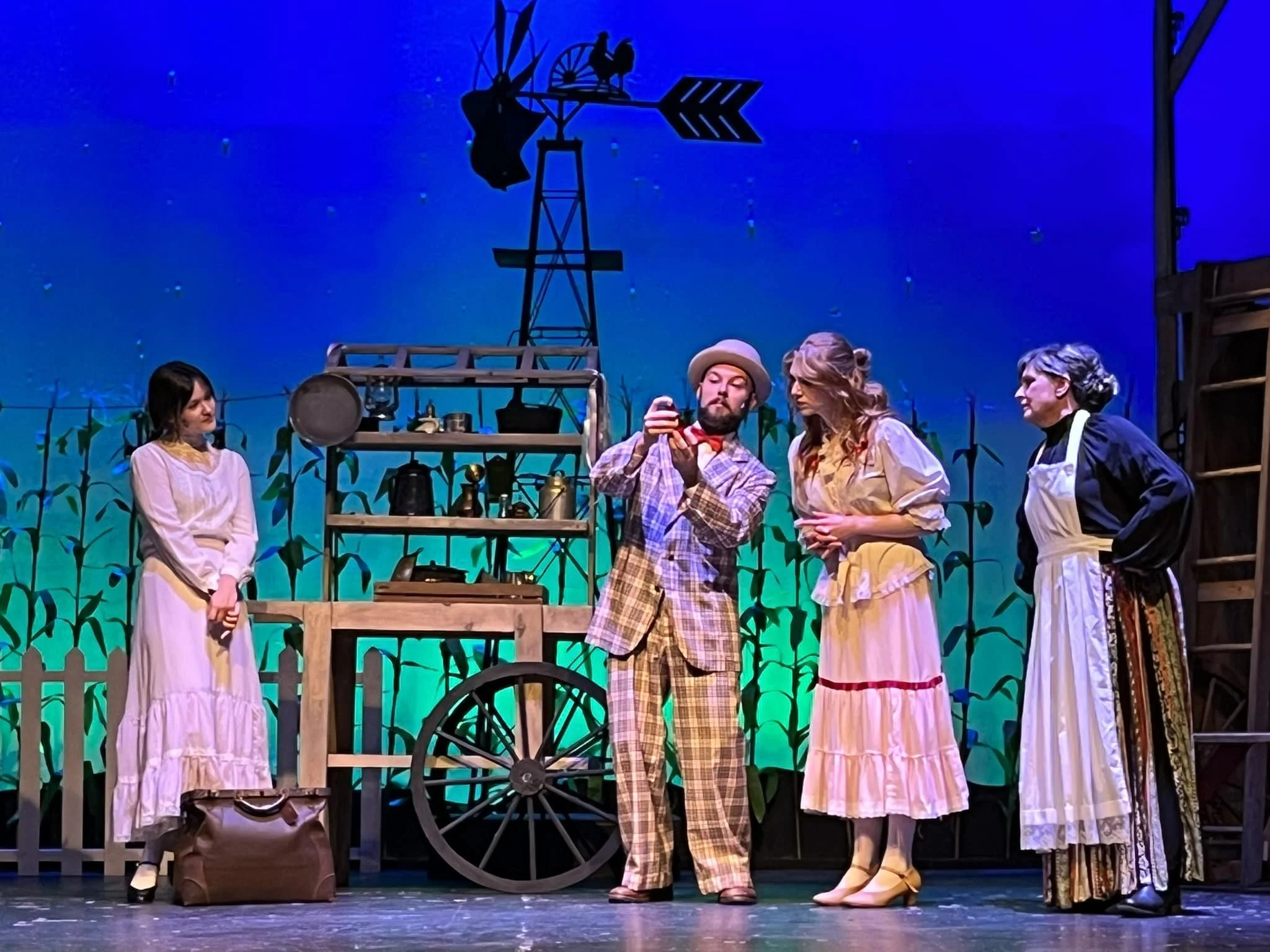 A theatrical stage scene with five actors in period costumes, a wooden cart, lantern, and a windmill prop, set against a starry night backdrop and a farm fence.