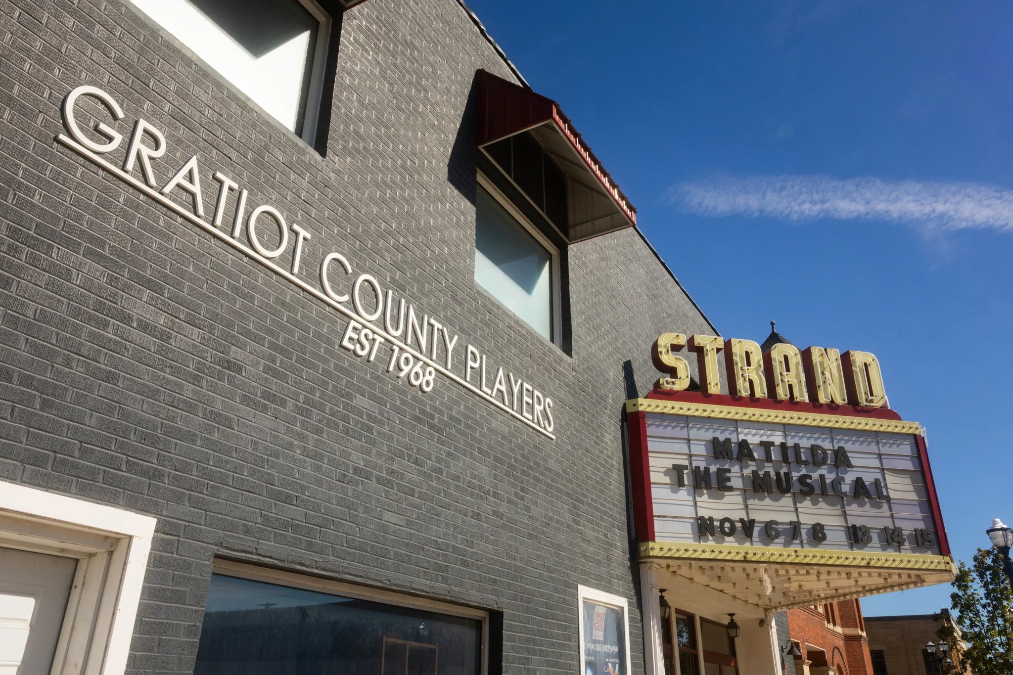 A horizontal photograph of the front of the Strand Theater in Alma Michigan. The building is gray brick with Gratiot County Players EST 1968 attached in white text. On the right it features a red and yellow marquee advertising Matilda The Musical.