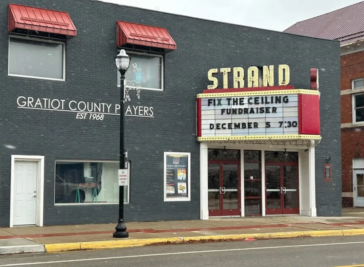 The exterior of a theater with a marquee that reads 'FIX THE CEILING FUNDRAISER' scheduled for December 5 at 7:30. The theater is called 'STRAND', with large yellow letters on top. To the left, there is a sign for the Gratiot County Payers, established in 1968, painted on the dark gray brick wall. A street lamp is standing in front of the building, and the sidewalk and street with a yellow-painted curb are visible.
