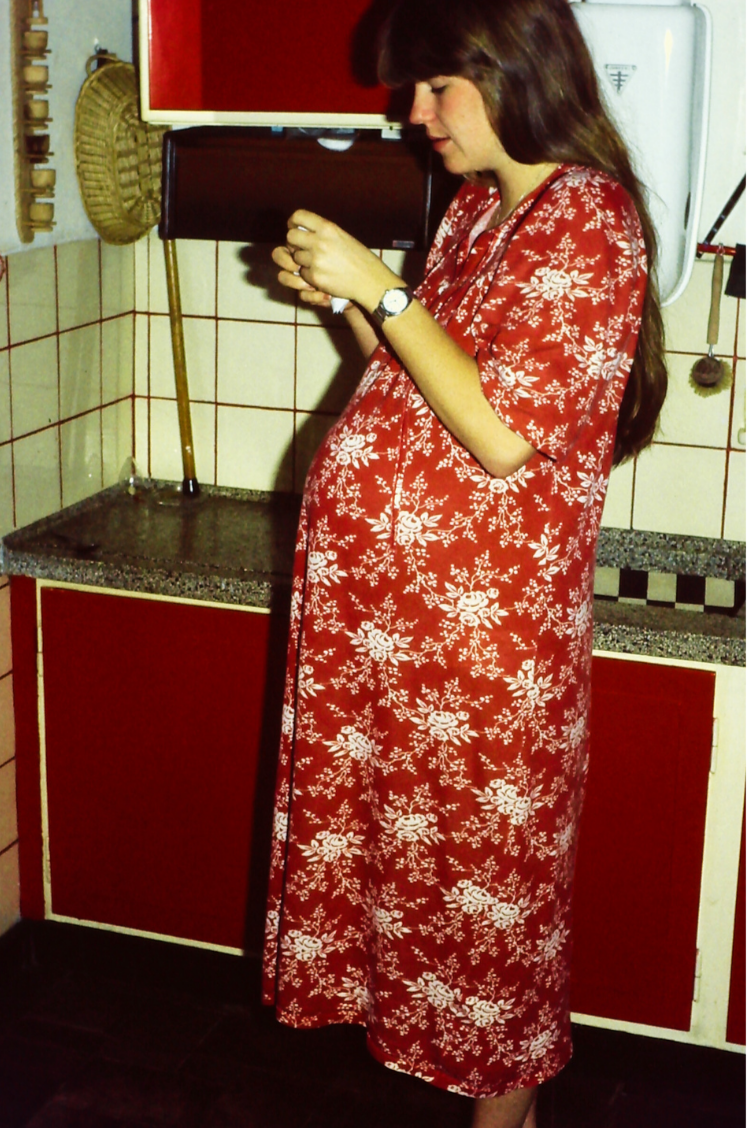 A woman with long dark hair in a red floral dress standing in a kitchen, looking at her phone.