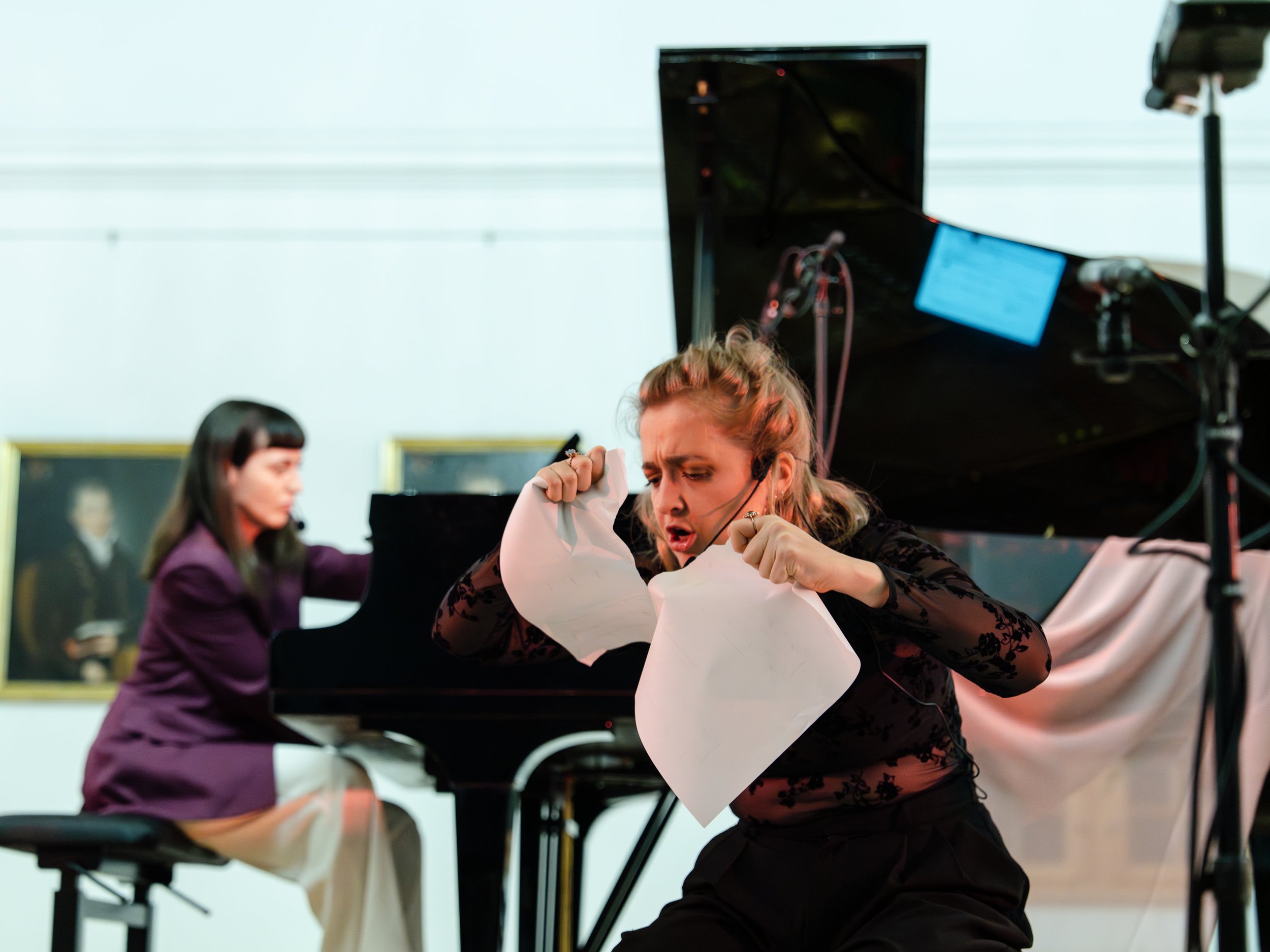 A woman in black with lace sleeves tearing a sheet of paper, with a woman playing piano in the background.