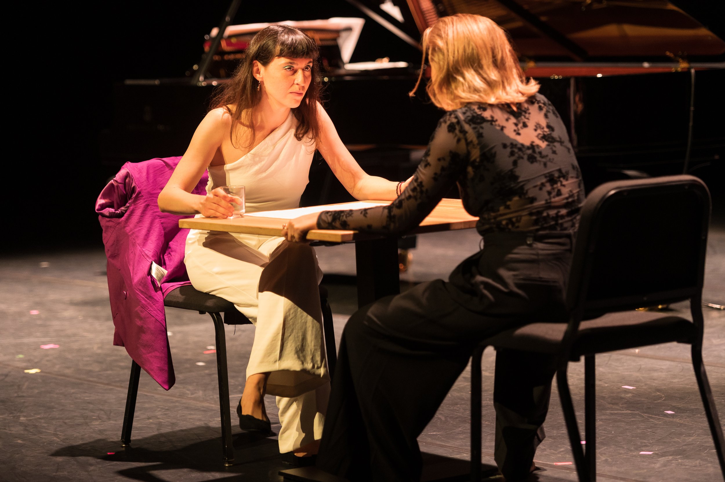Two women sitting at a table on a stage, engaged in conversation, with a grand piano in the background.