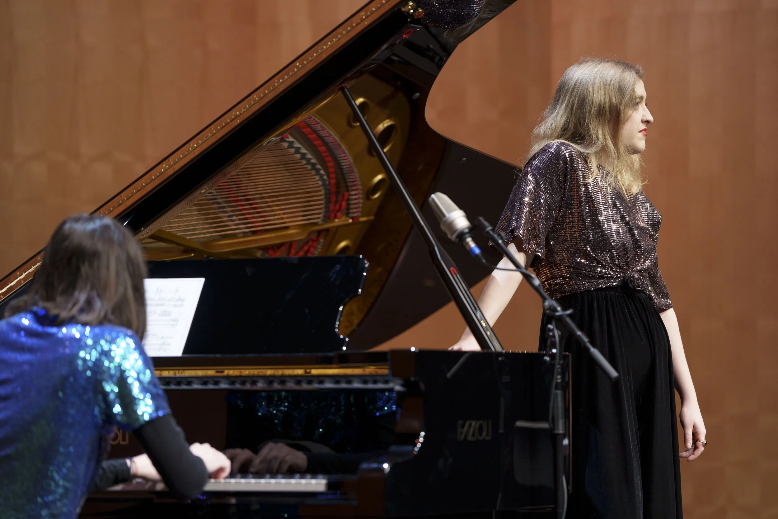 Two women performing on a stage with a grand piano. One woman is sitting at the piano with her back to the camera, wearing a sequined blue top. The other woman is standing beside the piano, facing to the right, wearing a shiny brown top and black pan
