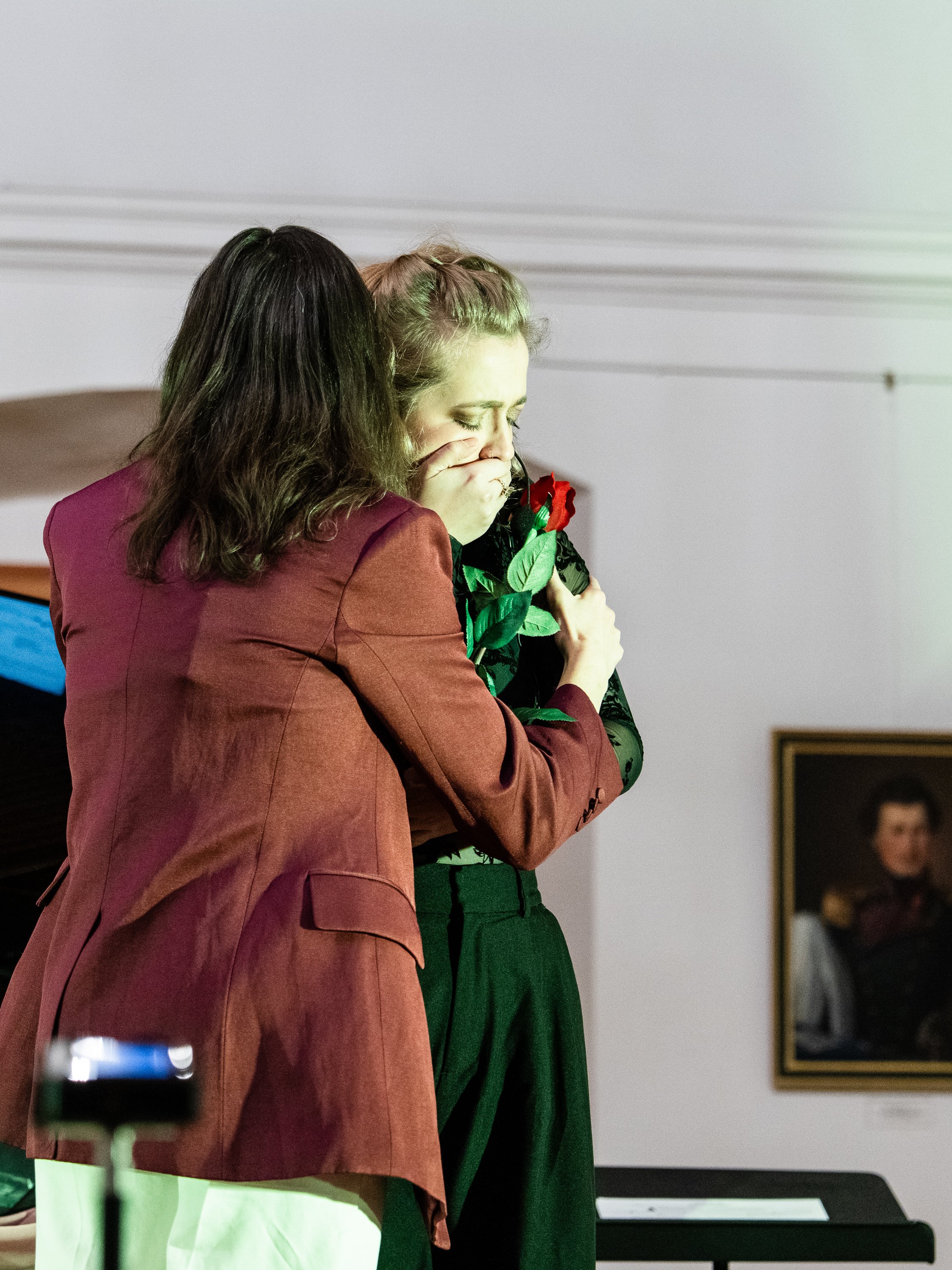 Two women hugging, one with her eyes closed and a tissue, holding a red rose, in an indoor setting with a portrait in the background.