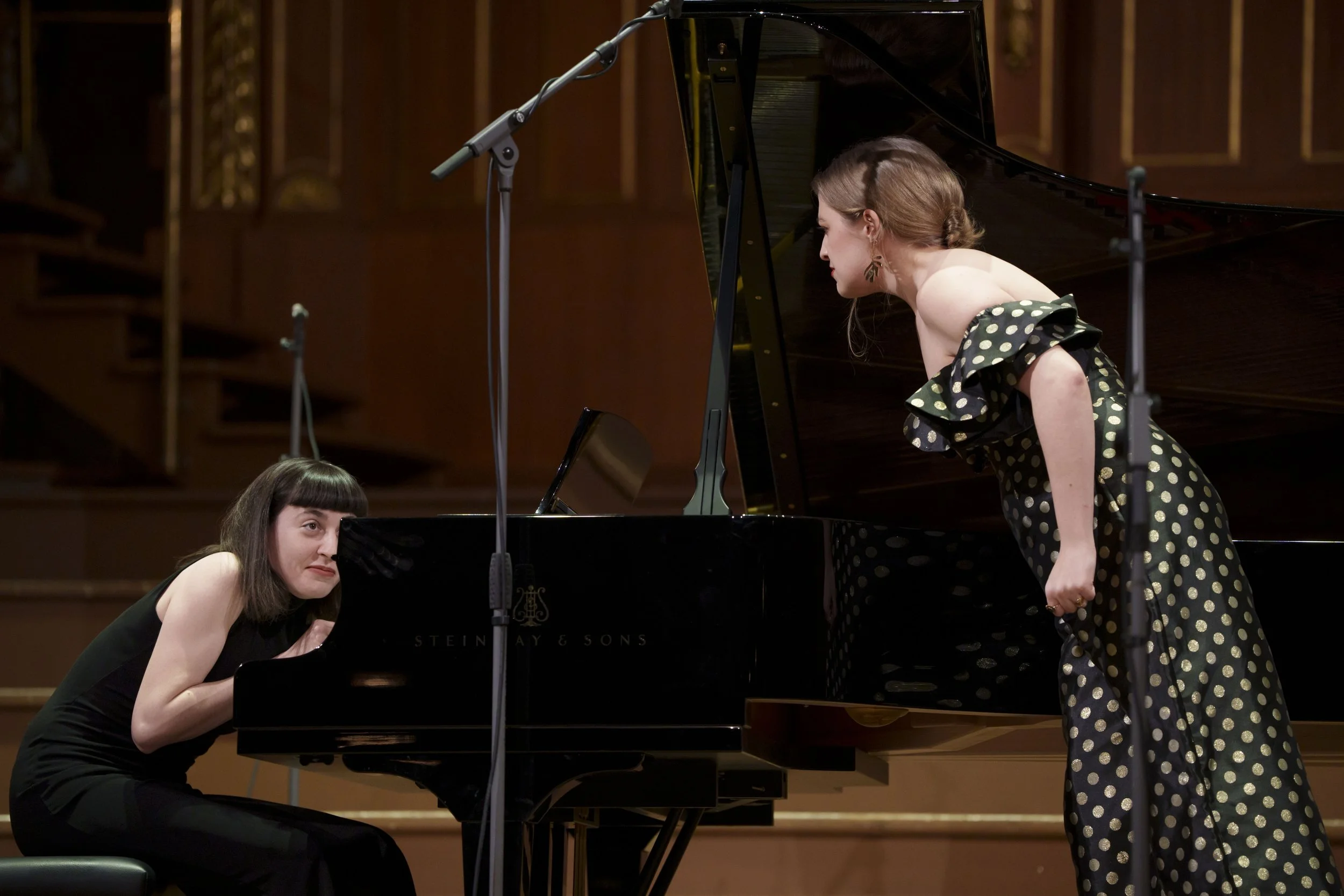 A woman in a black dress sitting at a grand piano, looking up at a woman in a polka dot dress, who is leaning towards her on stage during a musical performance.