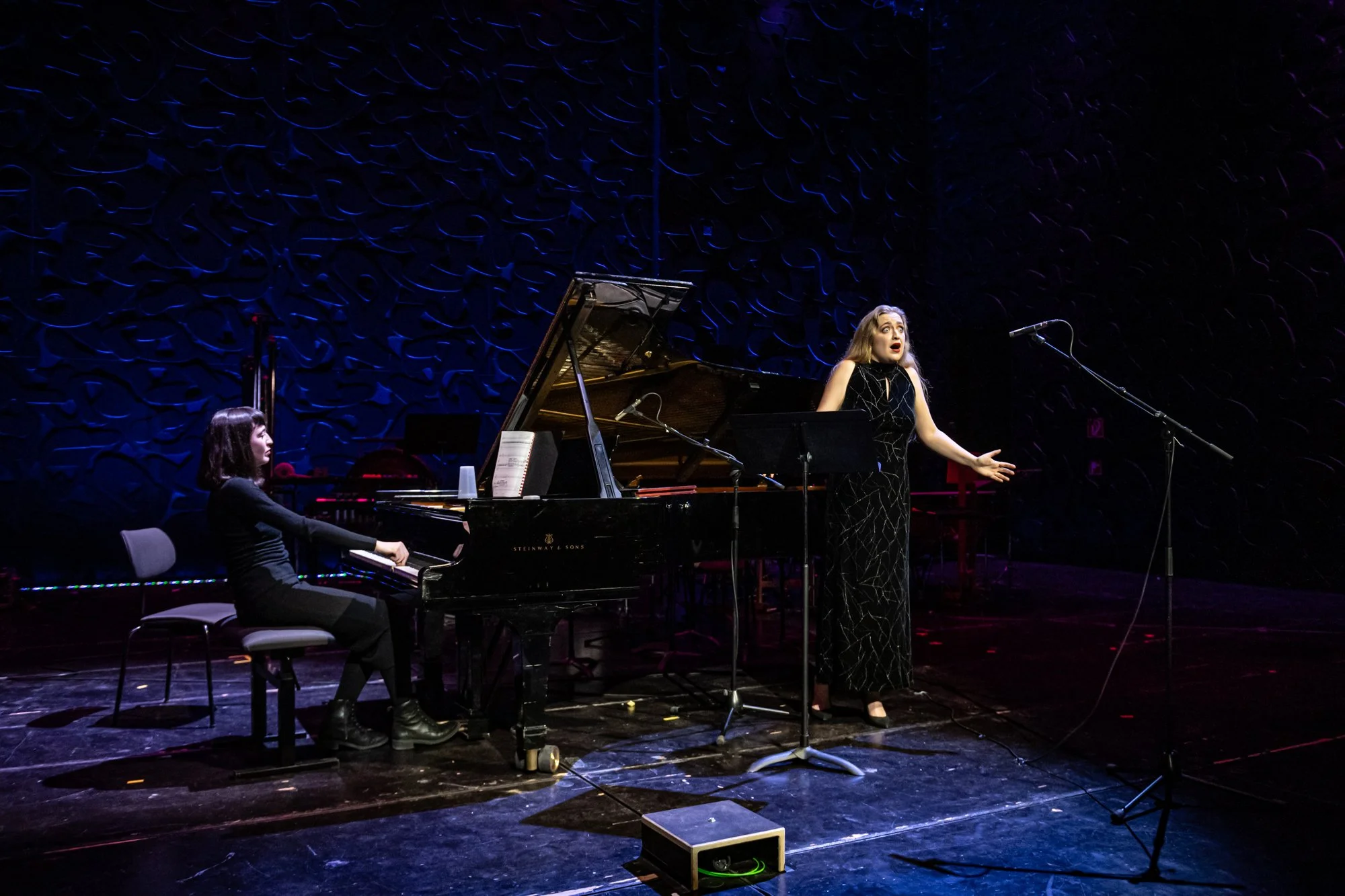 A woman singing on stage with a pianist accompanying her during a performance. The stage has a dark, textured background, with microphones set up in front of them.