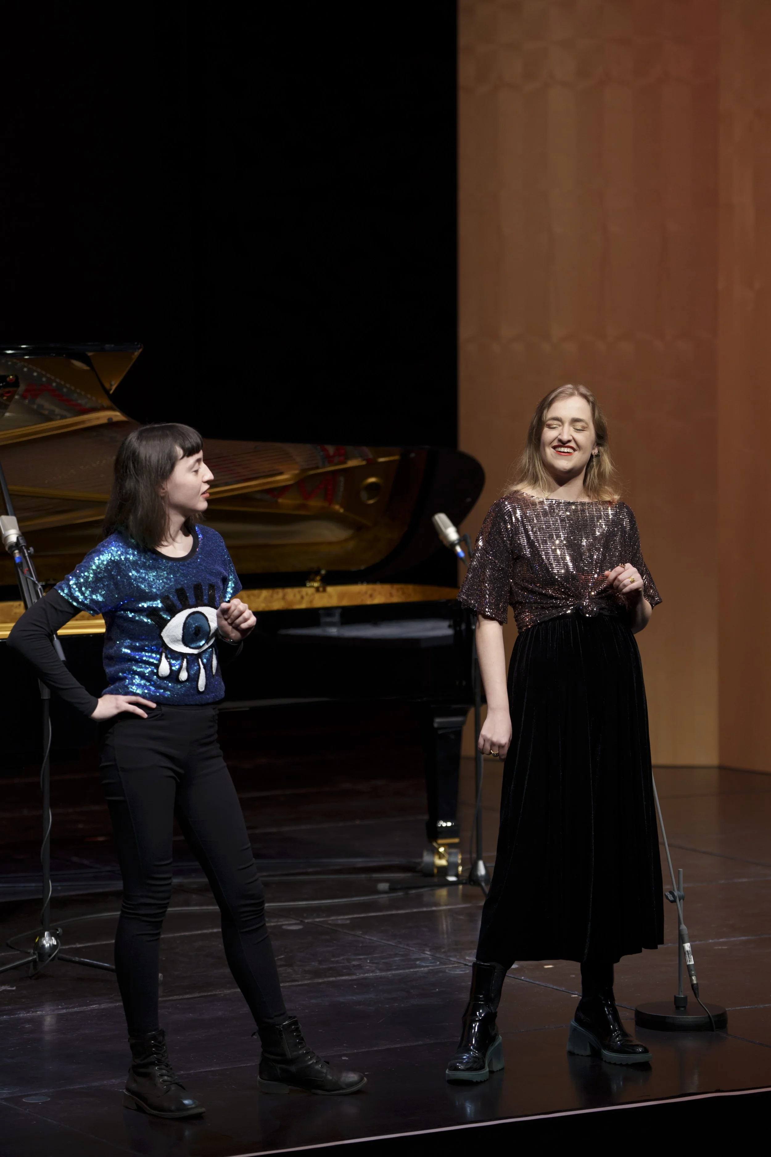 Two women stand on stage in front of a grand piano, one with a sequined top and the other with a graphic tee, smiling and possibly singing during a performance or rehearsal.