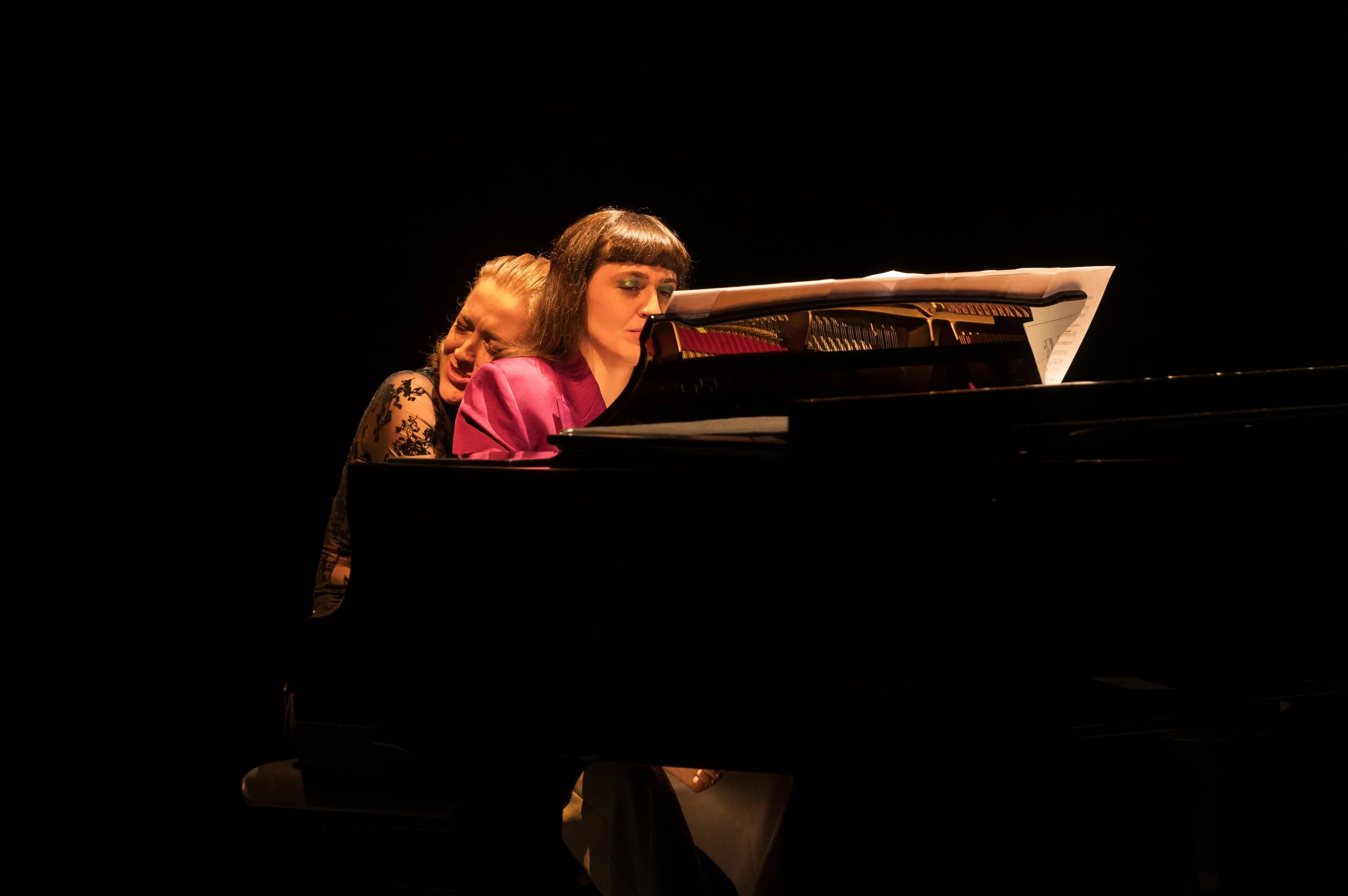 Two women playing the piano on stage; one in a black lace dress singing with her eyes closed, the other in a pink blazer looking at sheet music