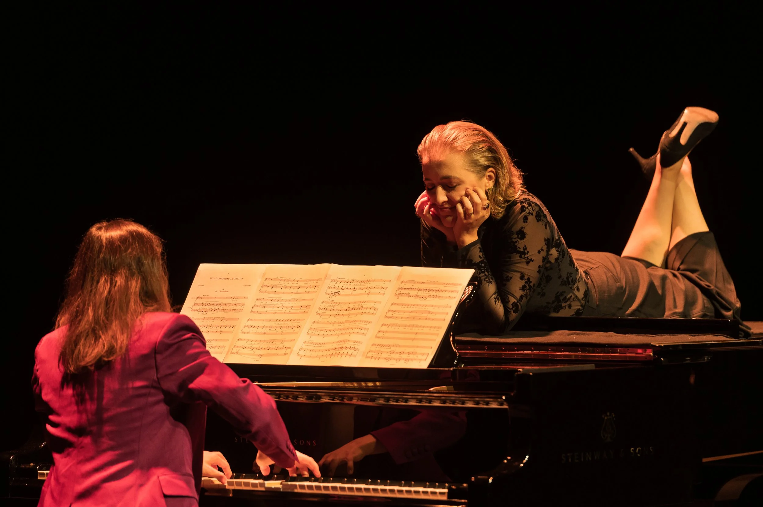Two women are performing a piano duet on stage, one sitting at the piano and the other lying on top of it, both looking at sheet music with a joyful expression.