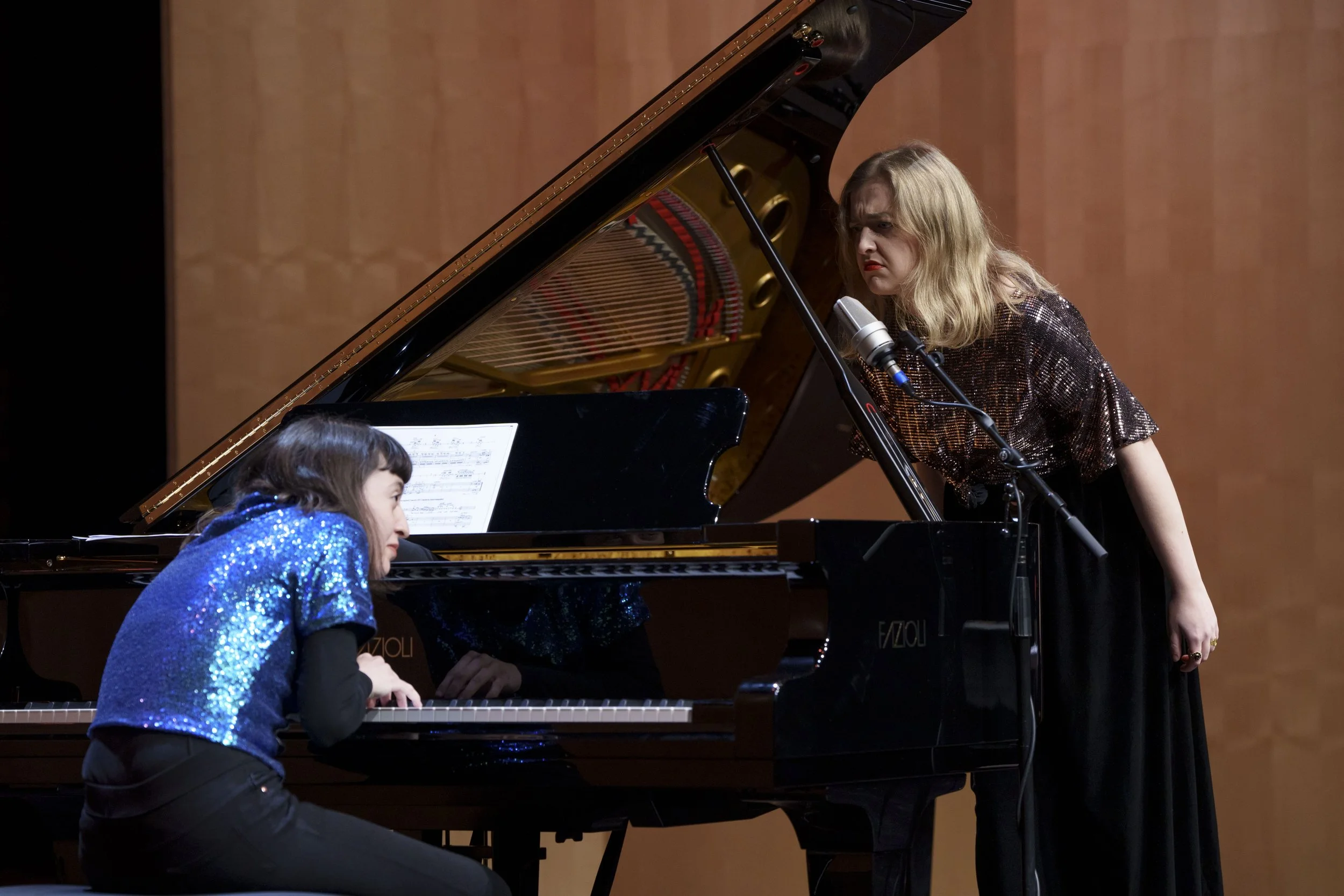 A woman with blonde hair in a shiny dark top is standing near a grand piano, singing into a microphone, while a young girl with dark hair wearing a sparkly blue top is sitting at the piano, playing and looking at sheet music.