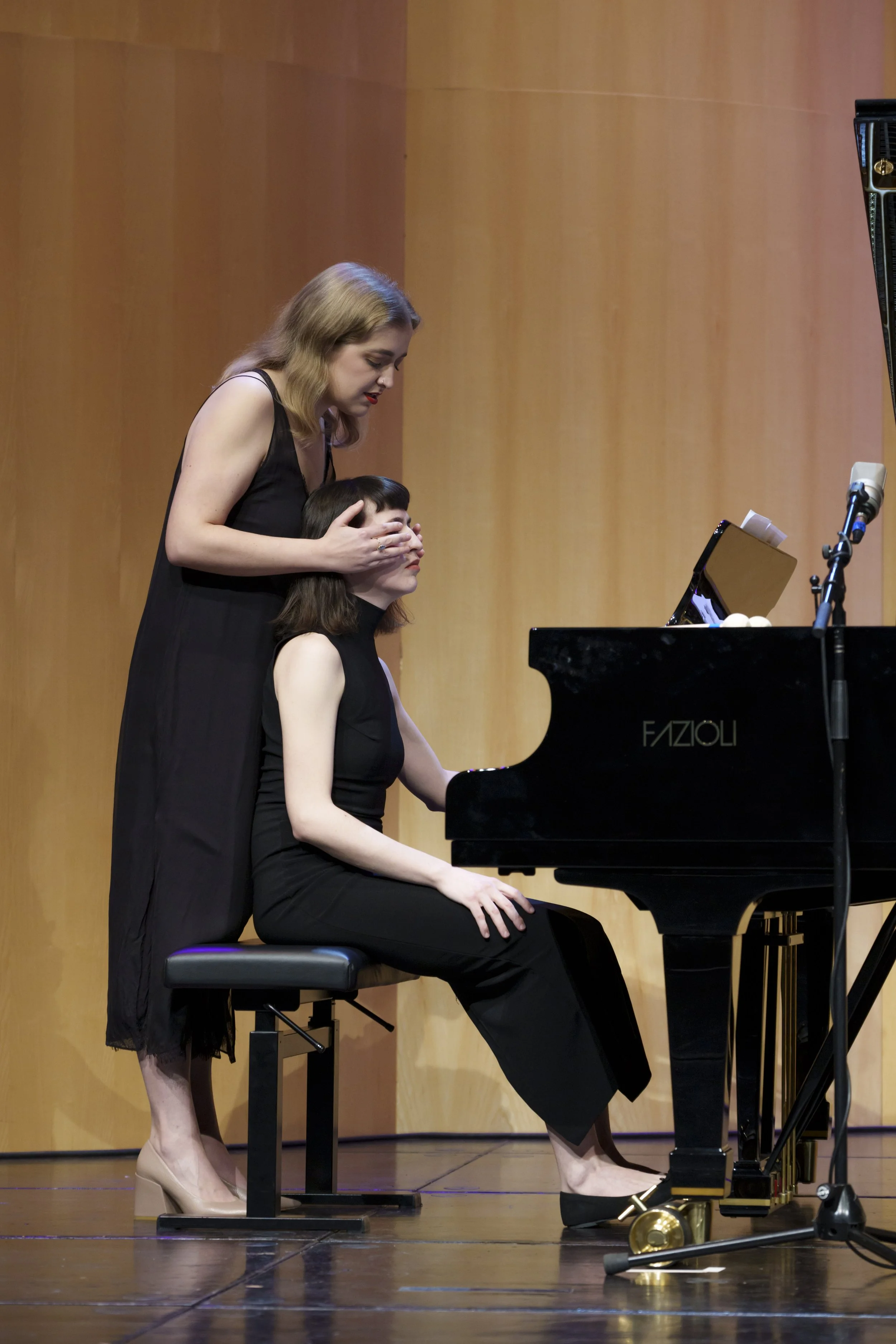 Two women on stage; one is seated at a piano, the other standing behind and holding her face.