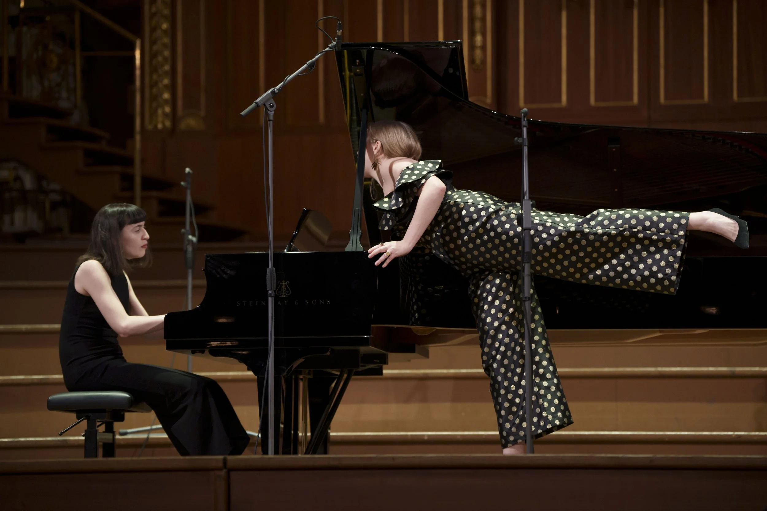 Two women performing on a grand piano on stage in a concert hall, one sitting at the piano and the other leaning on it, singing into a microphone.