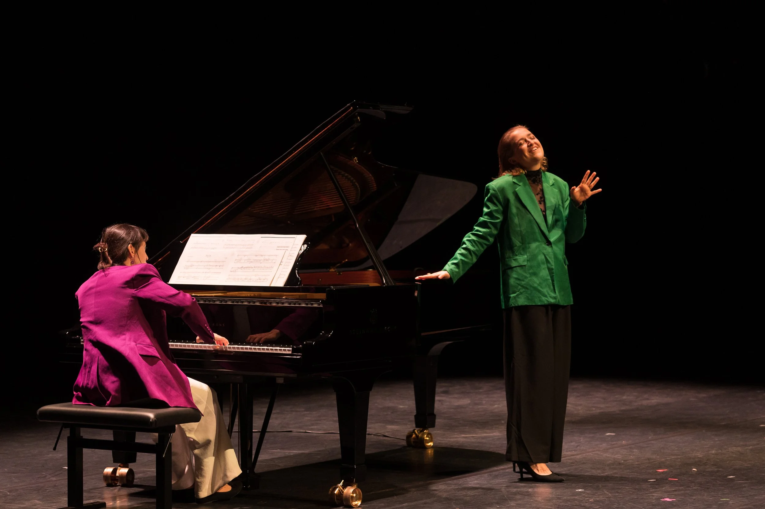 A woman in a green blazer and black pants is singing on stage with her eyes closed and her hand raised, while a young girl in a purple jacket plays a grand piano next to her.