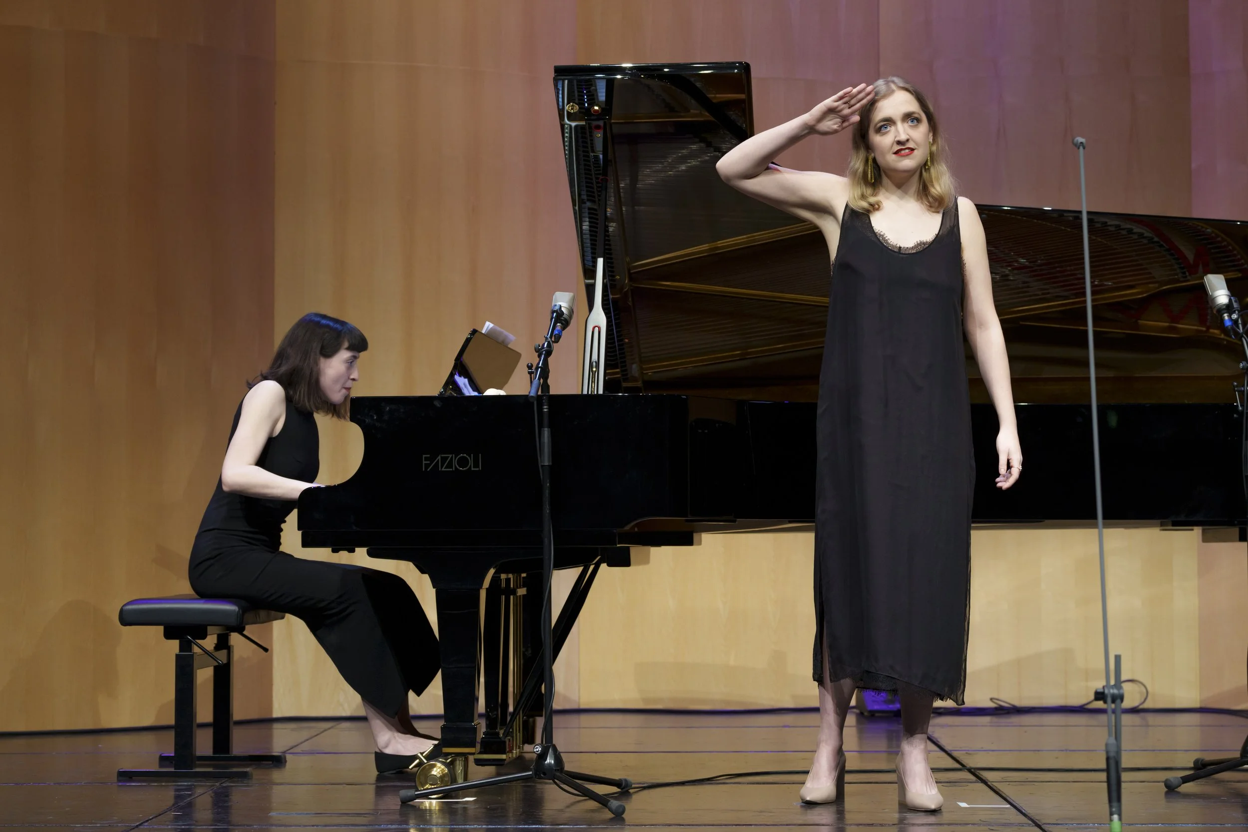 A woman saluting on stage next to a pianist playing a grand piano.