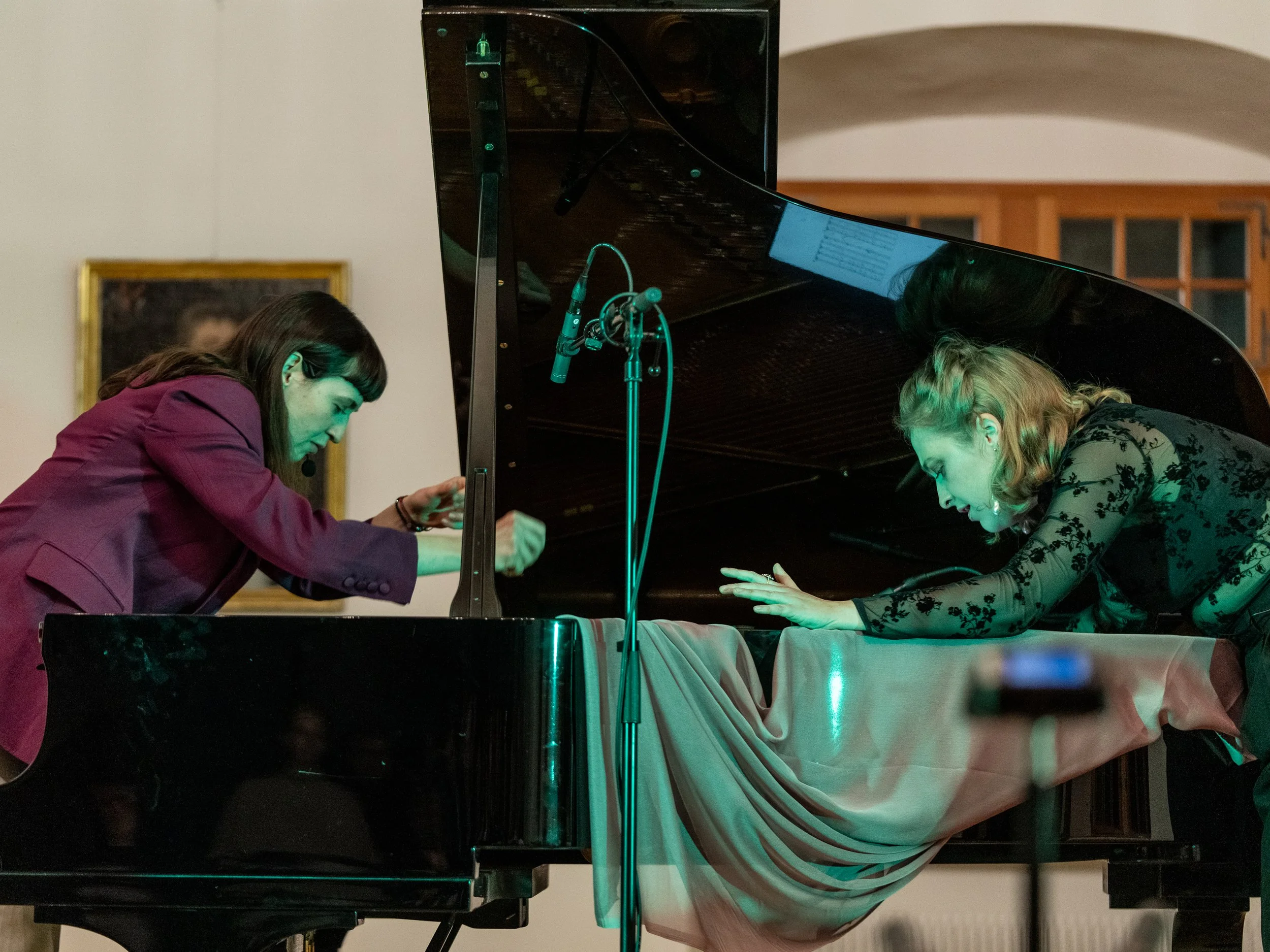 Two women are performing a piano duet, leaning forward with their hands on the piano, looking intently at the keys, in a room with a painting on the wall.