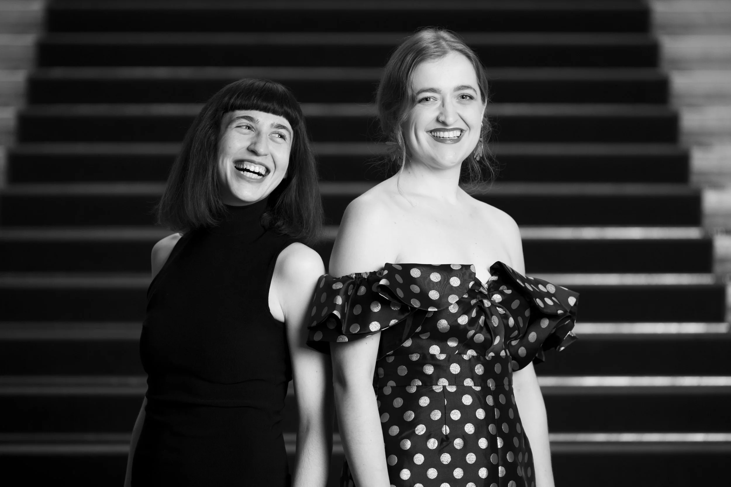 Two smiling women in black and white standing on stairs, one wearing a sleeveless black dress and the other in an off-shoulder polka dot dress.