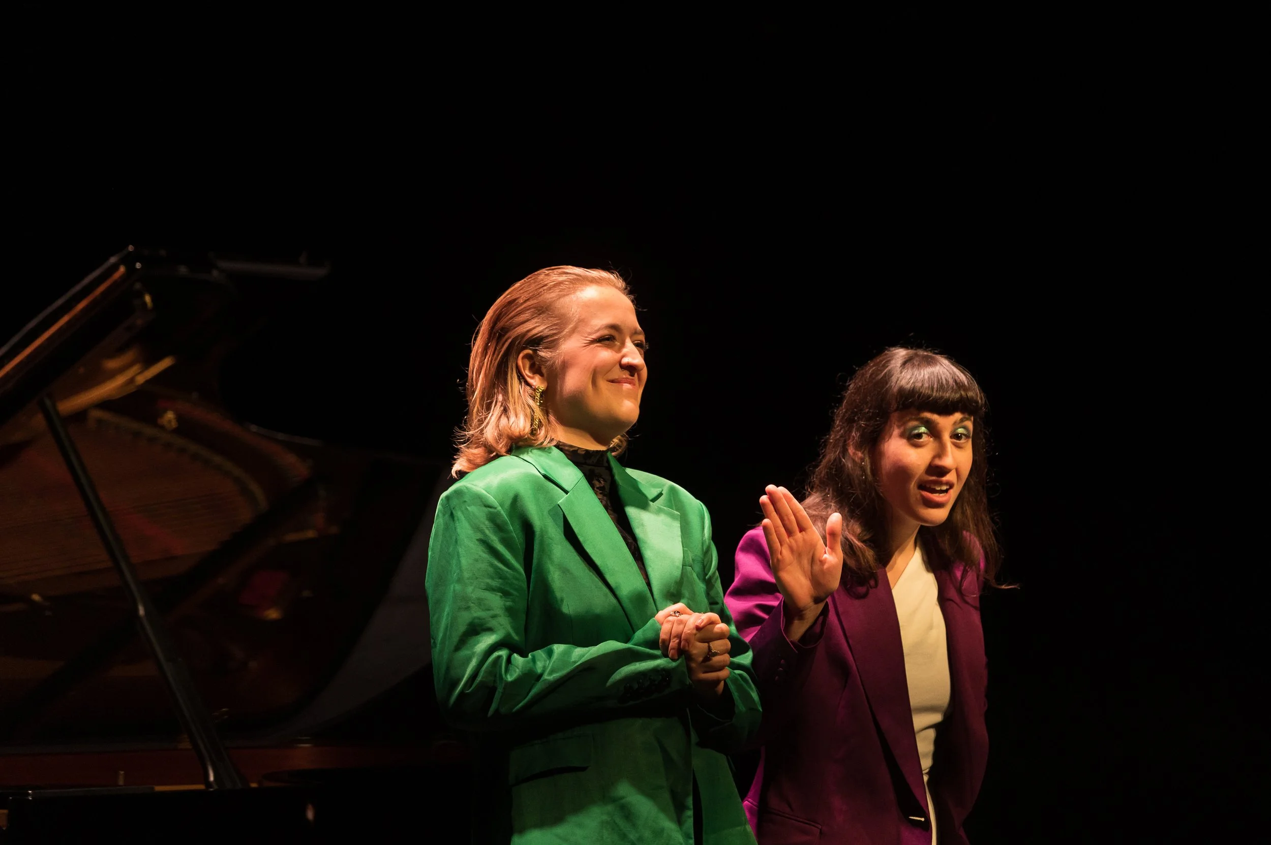Two women standing together on stage, with a grand piano behind them, one waving and smiling, dressed in colorful blazers, against a black background.
