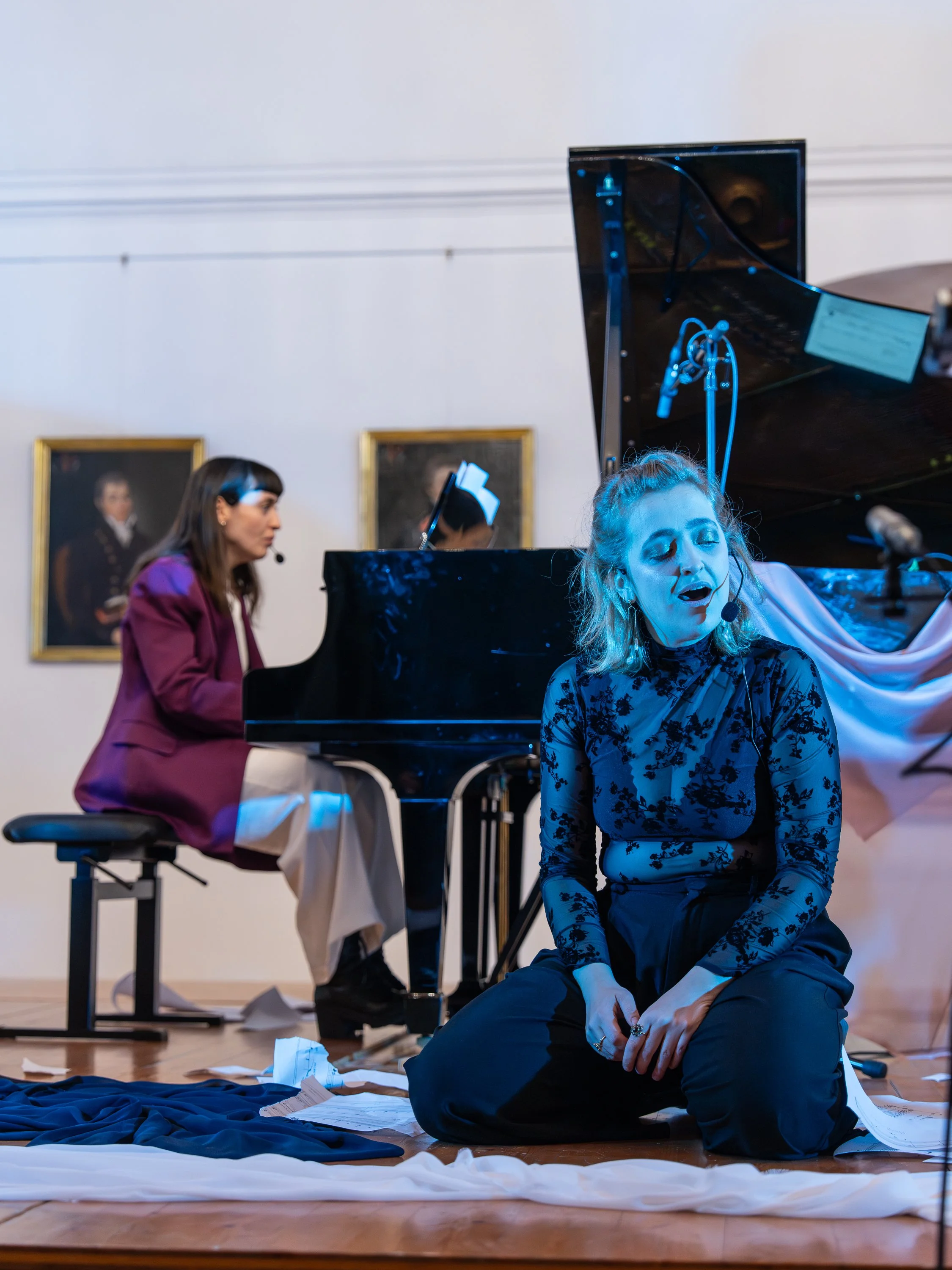 A woman singing with a headset microphone on stage, kneeling on the floor with papers around her, and a woman playing the piano in the background.