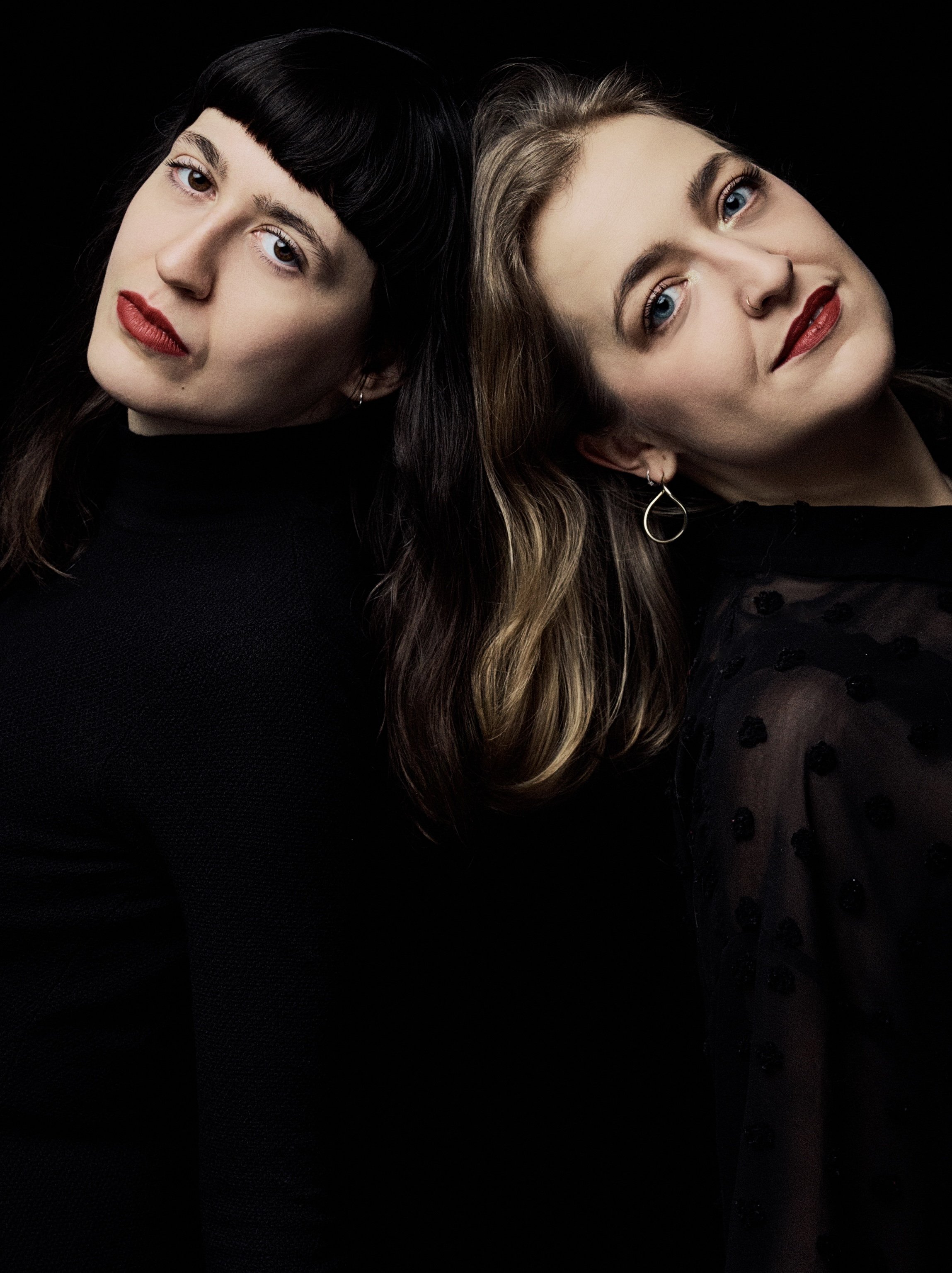 Two women with contrasting hair colors and styles, posing against a black background. The woman on the left has straight, dark hair with bangs and wears a black top. The woman on the right has wavy, light brown hair with blonde highlights, wearing a 