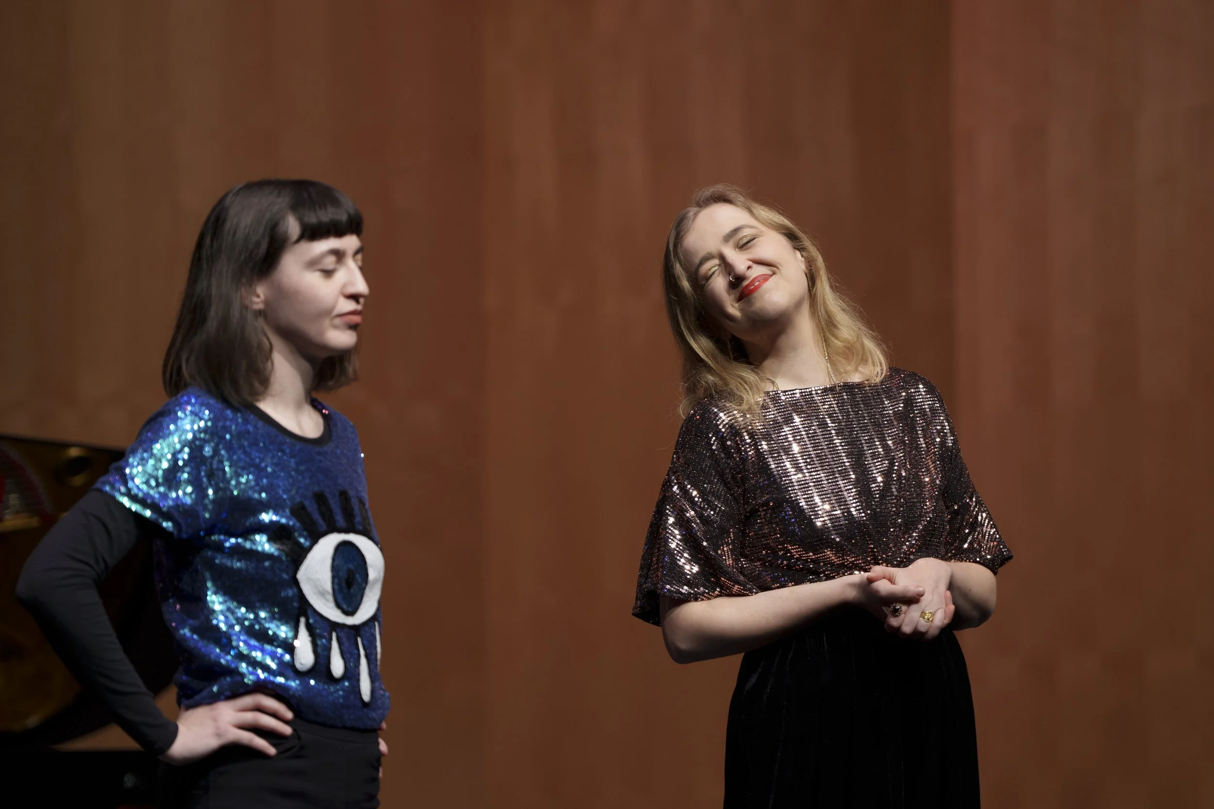 Two women standing on a stage with wooden background, one with dark hair wearing a blue sequin top with an eye symbol, the other with blonde hair wearing a shiny bronze sequin top, both smiling.