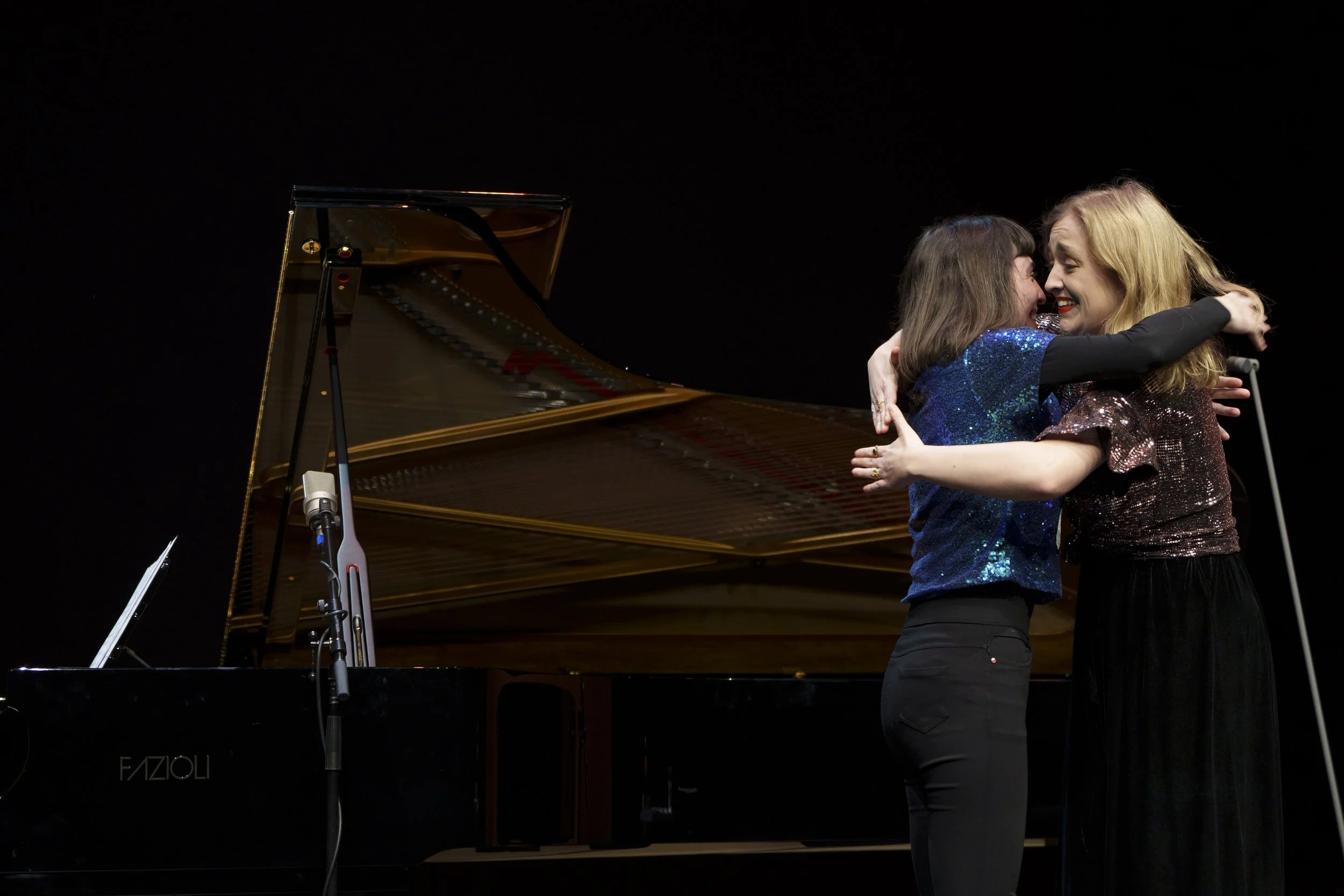 Two women embrace and smile on stage in front of a grand piano, with the woman on the right holding a microphone.